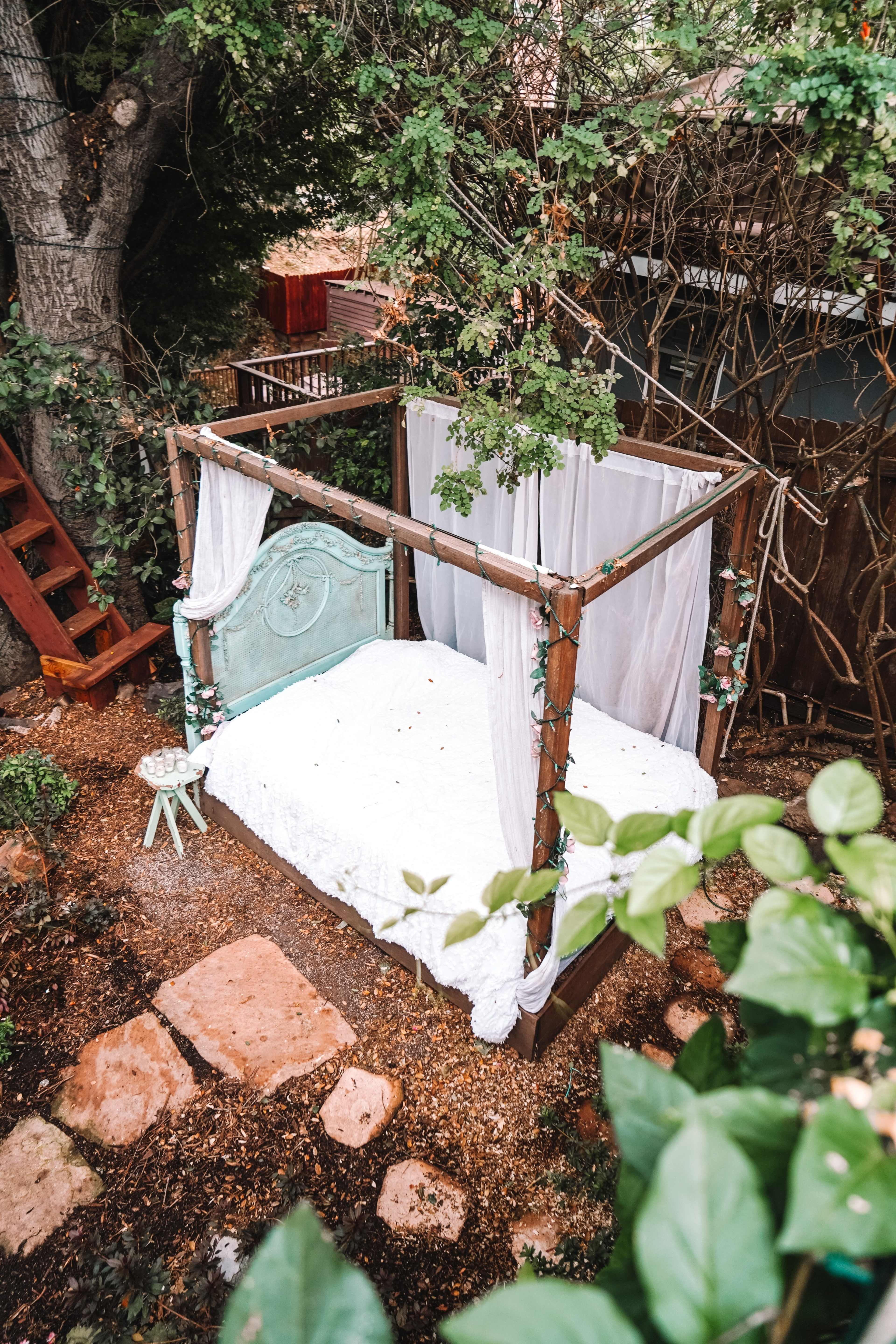 A canopy bed with a light-colored mattress is situated in a secluded outdoor area surrounded by trees and greenery.