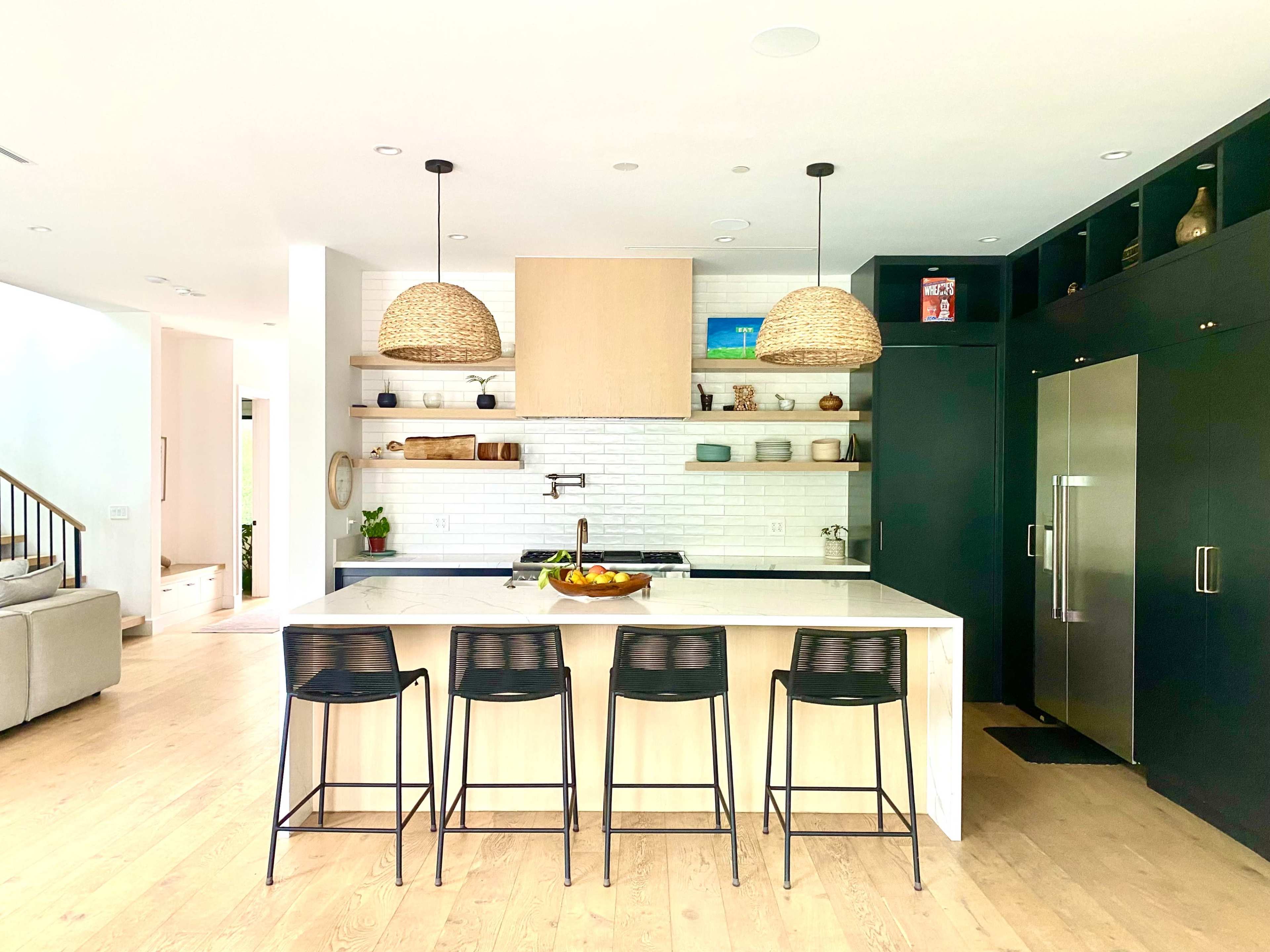 A modern kitchen features a large island with four black bar stools, two woven pendant lights, and open shelving displaying various kitchen items against a white tile backsplash.