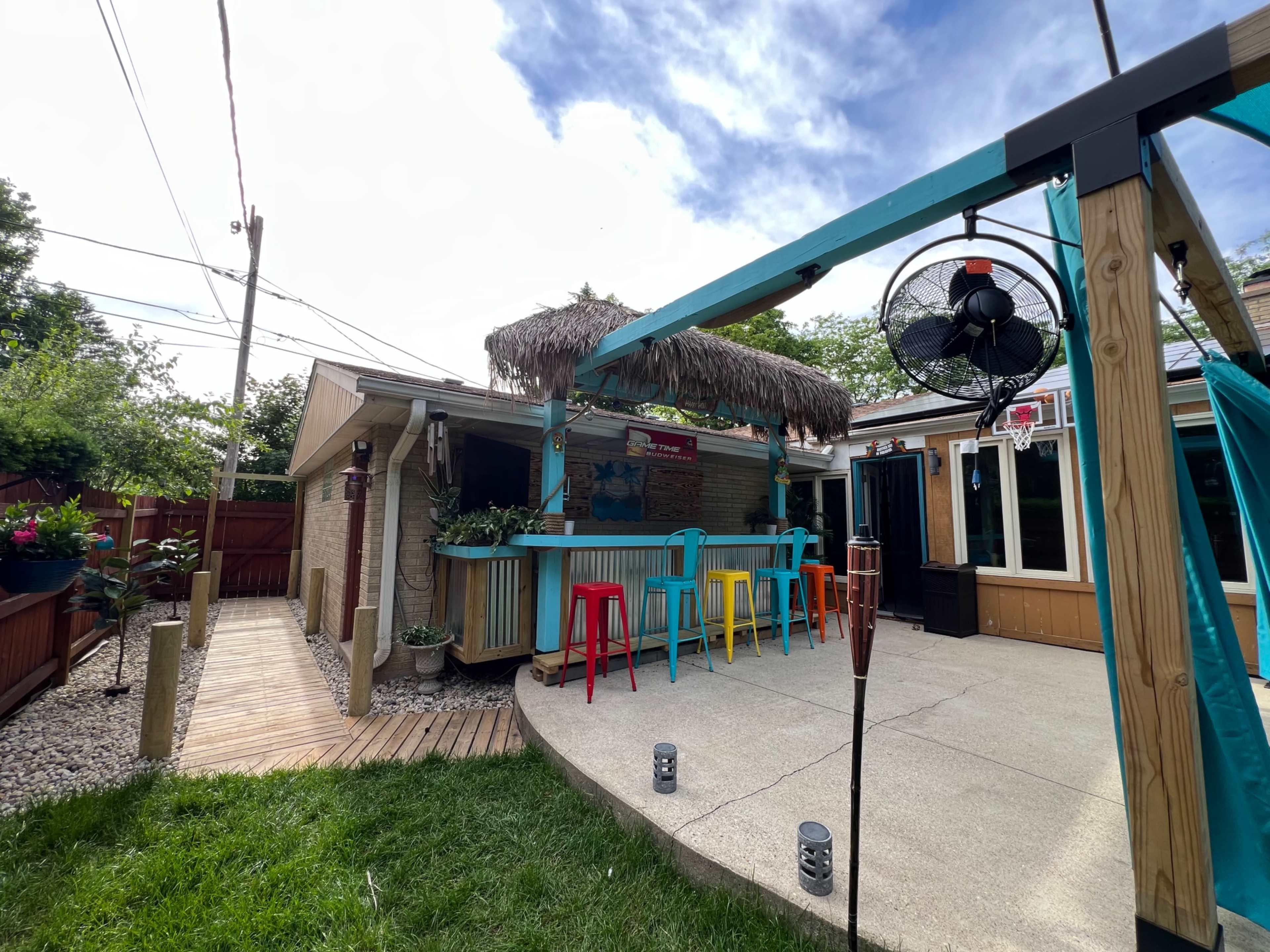 A backyard area features a bar with colorful stools, a thatched roof, and a patio space surrounded by greenery and wooden fencing.