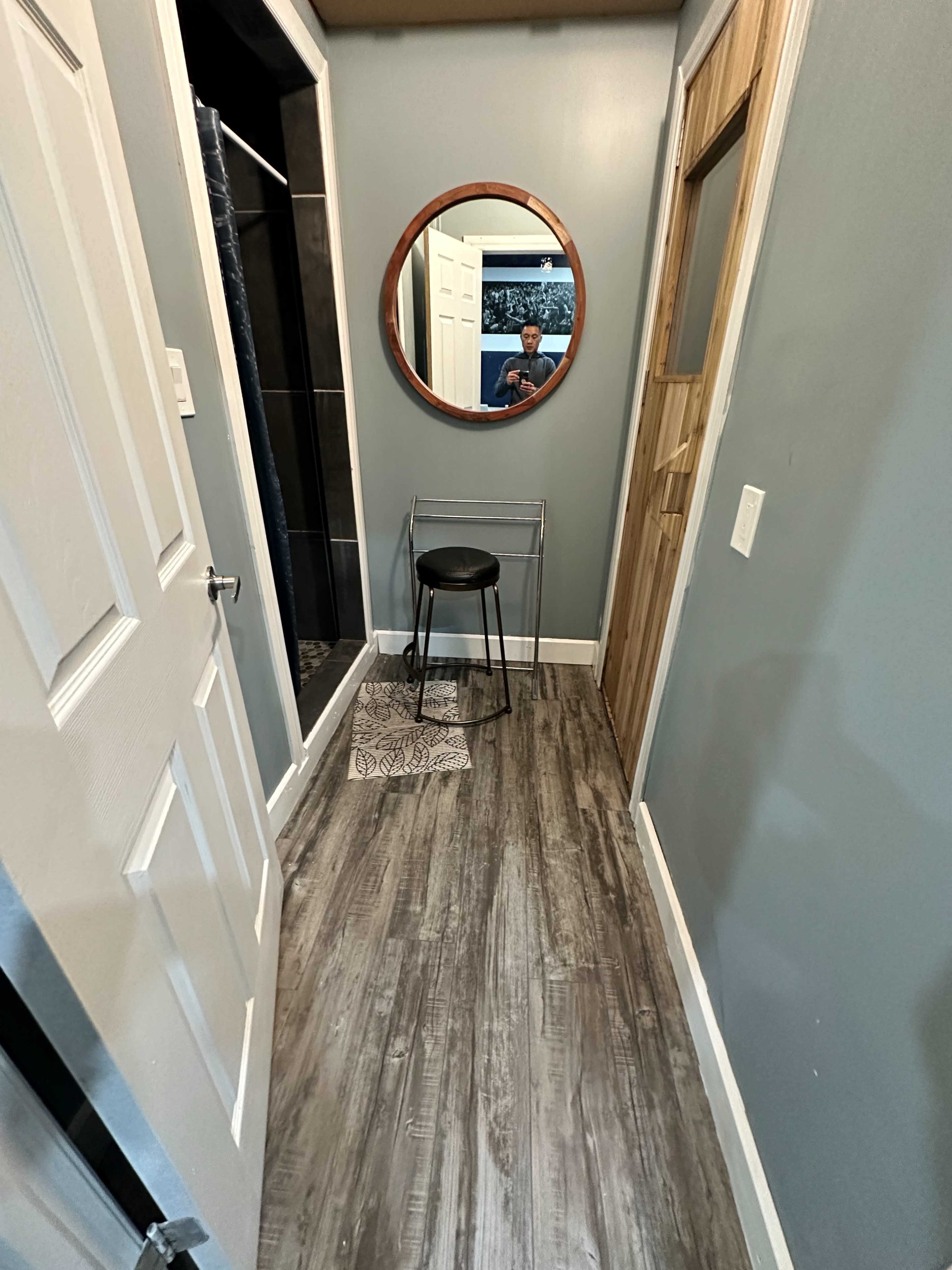 The image shows a narrow hallway with a round mirror mounted on the wall, a black stool, and a door leading to a bathroom area.