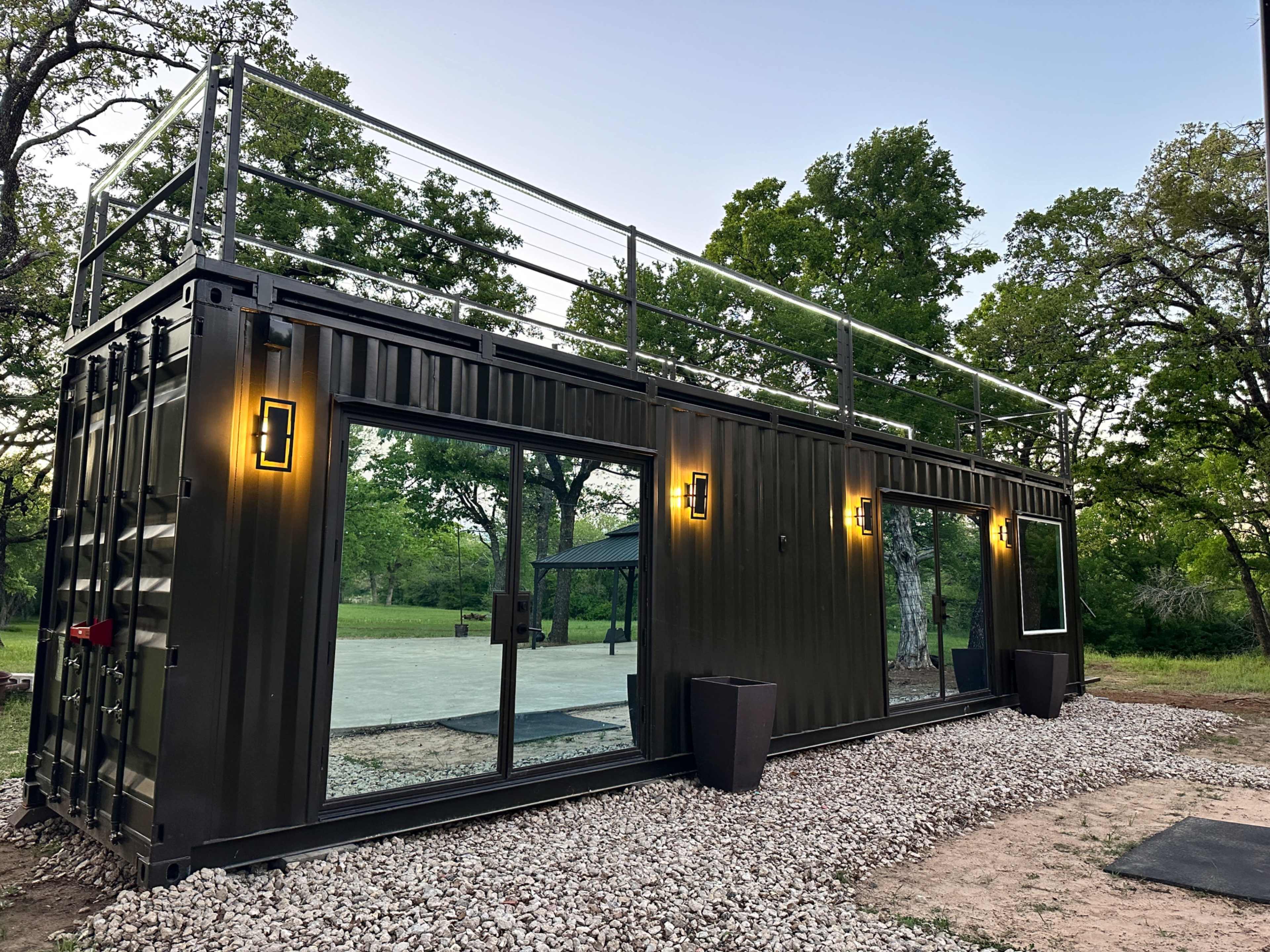 The image shows a modern shipping container home with large windows and a rooftop terrace, set in a grassy area surrounded by trees.