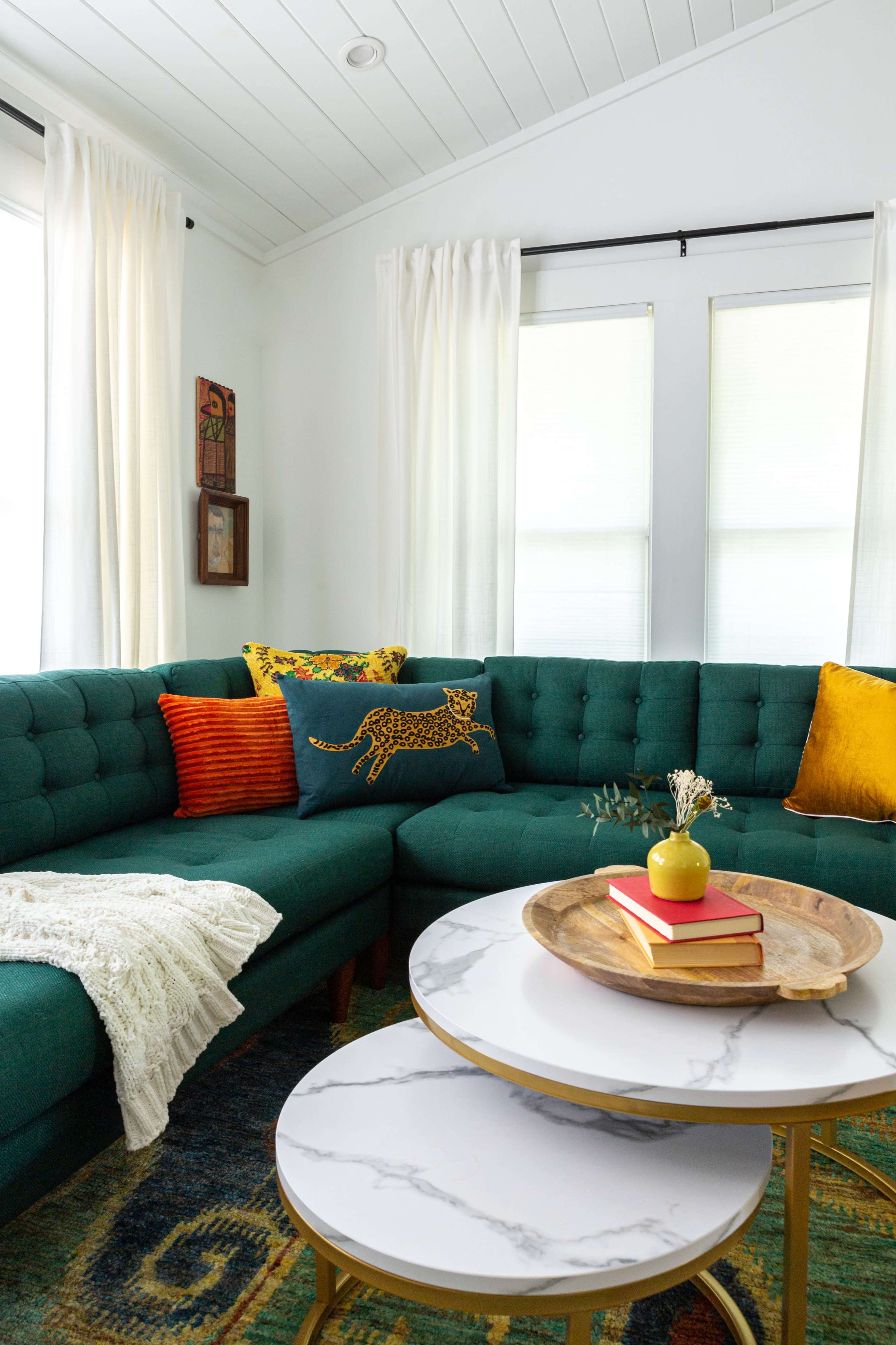 A green sectional sofa with decorative pillows is arranged around a marble-top coffee table in a bright, modern living room.