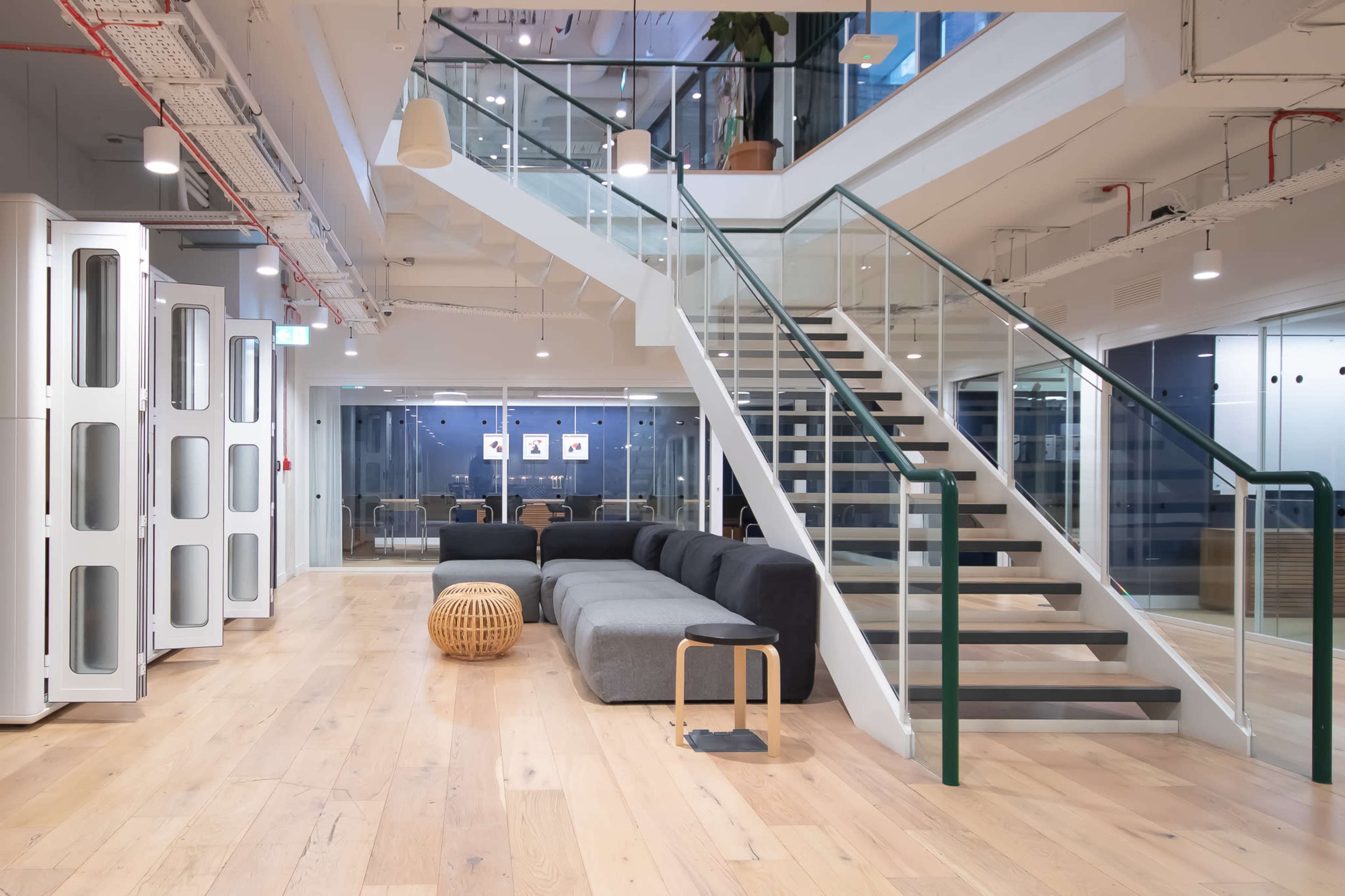 The image shows a modern office interior featuring a staircase, a gray sectional sofa, wooden flooring, and glass partitions.