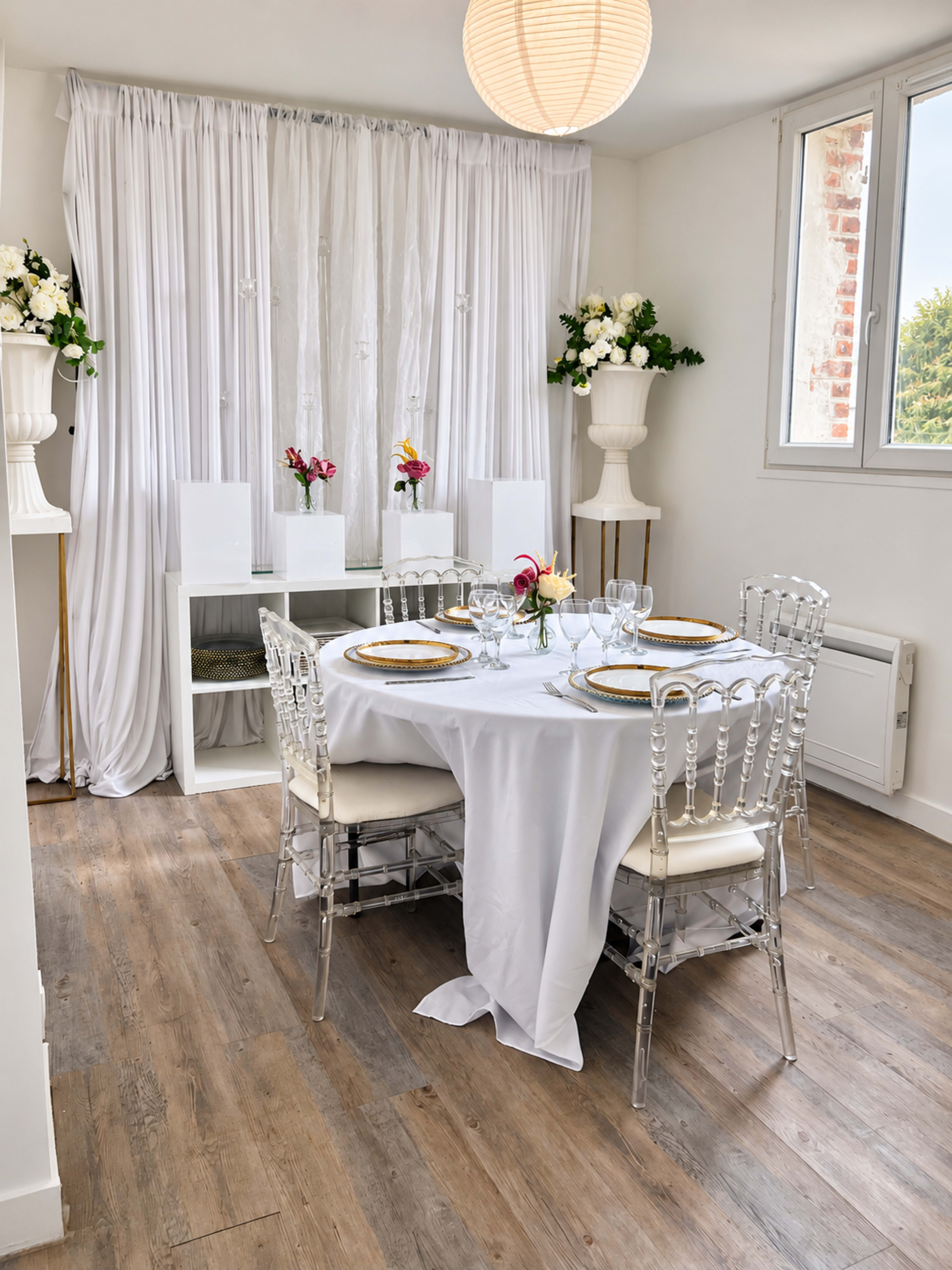 A round dining table is set with white tablecloth and clear chairs, surrounded by white curtains and potted flowers in a bright, modern room.
