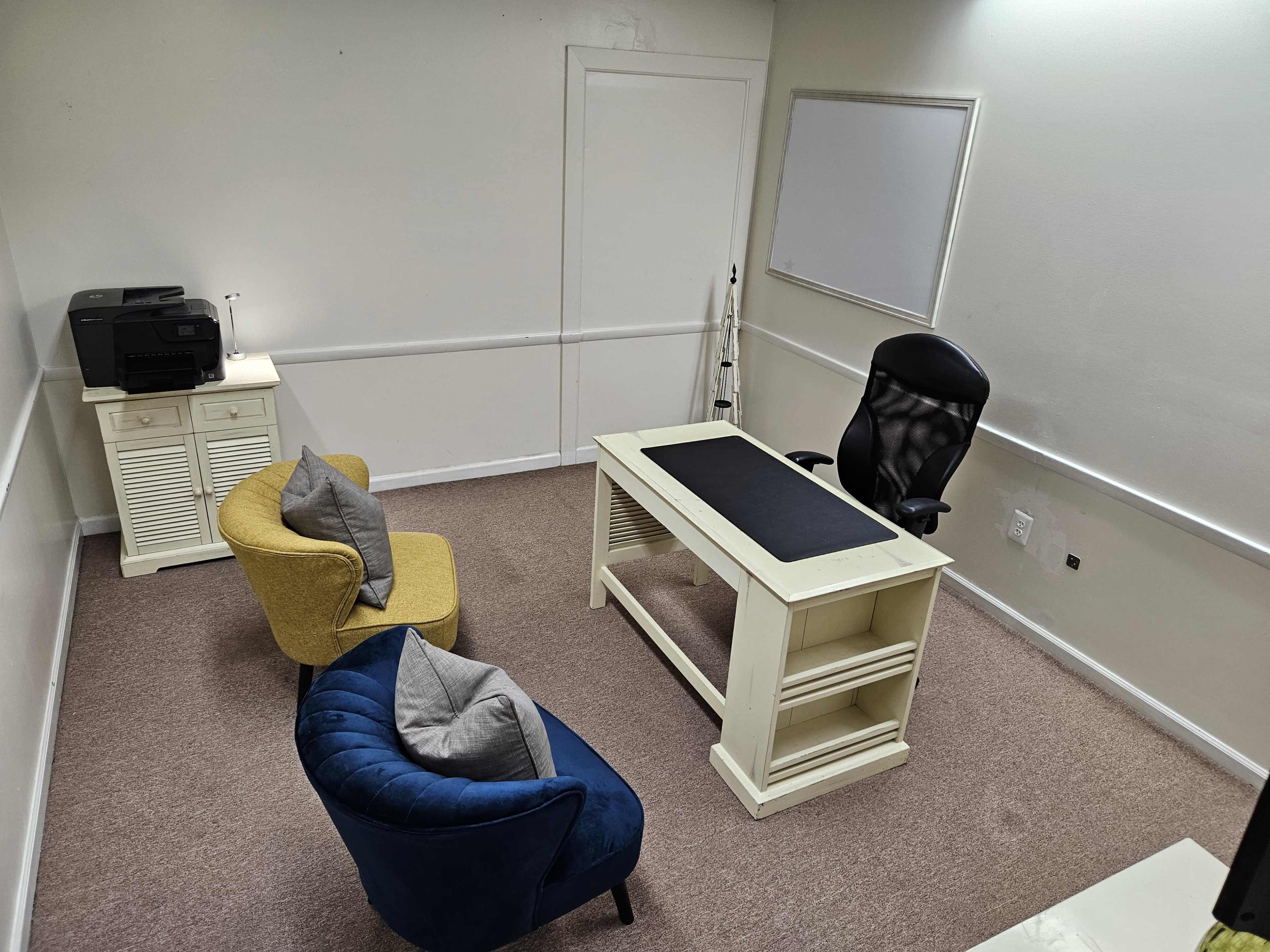 The image shows a small office space with a white desk, a black office chair, two armchairs, a printer, and a whiteboard on the wall.
