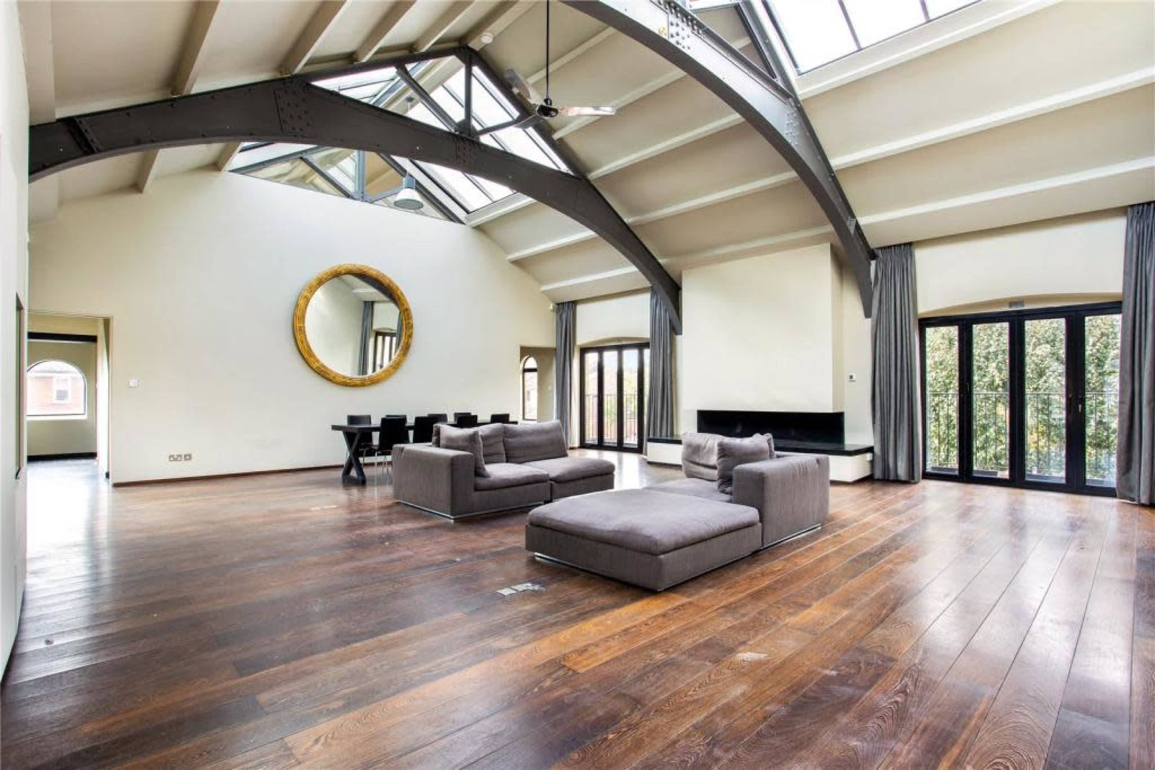 A spacious living room features a high ceiling with skylights, two gray sofas, a round mirror, and large glass doors leading to an outdoor area.