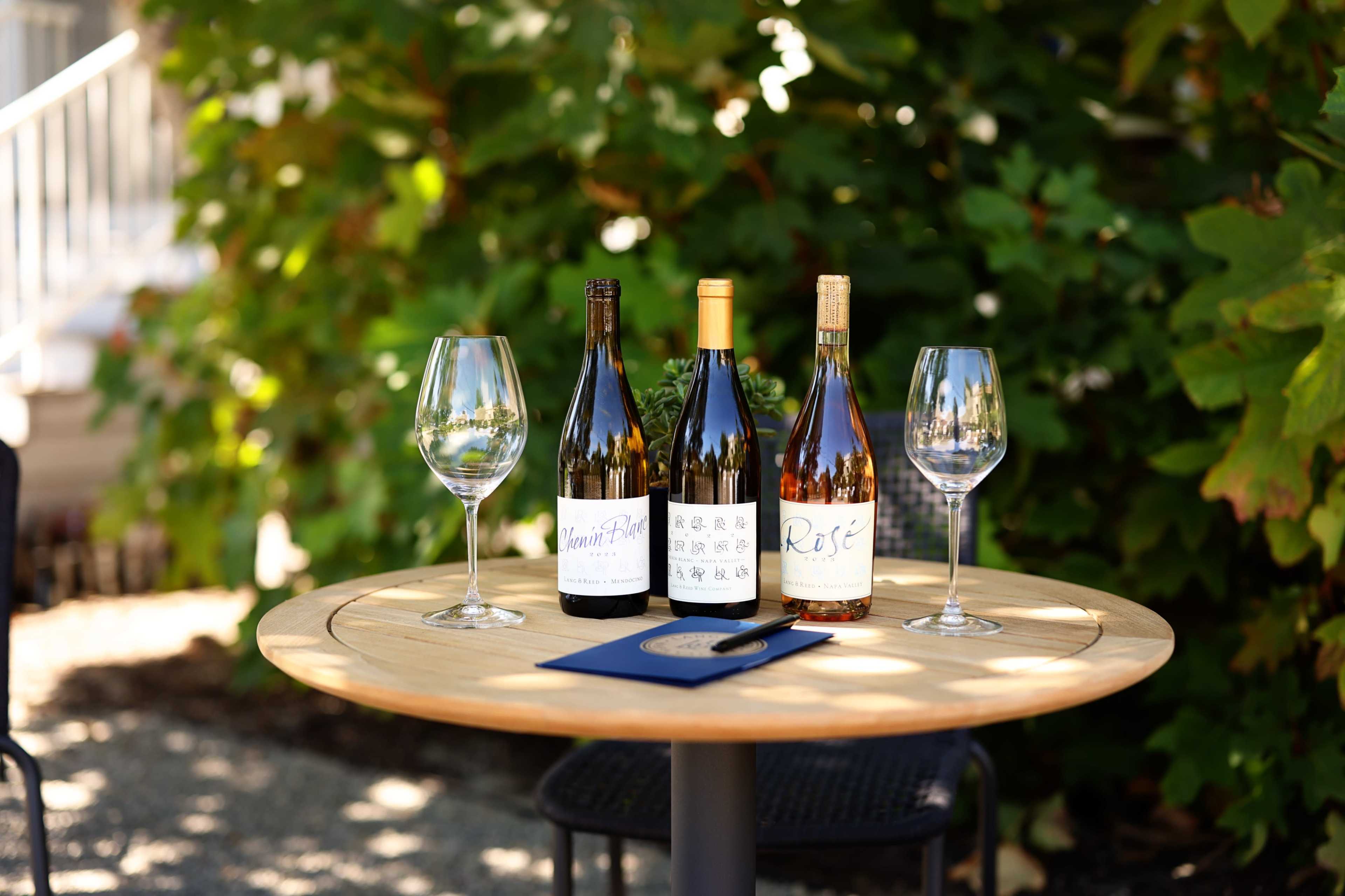 A wooden table outdoors displays three bottles of wine, each with a distinct label, accompanied by two empty wine glasses and a menu.