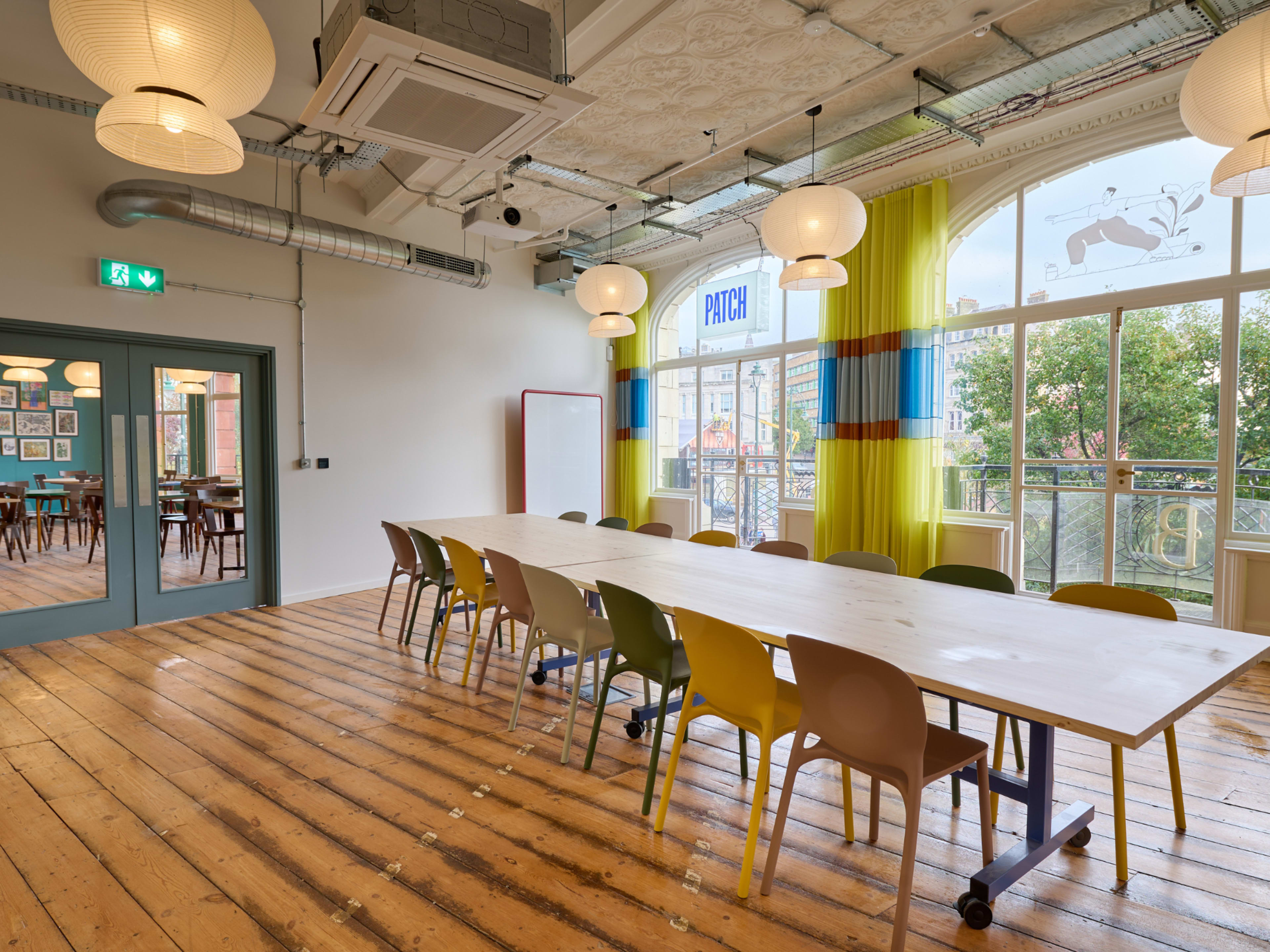 The image shows a spacious room with wooden floors, a long table surrounded by colorful chairs, and large windows adorned with sheer curtains.