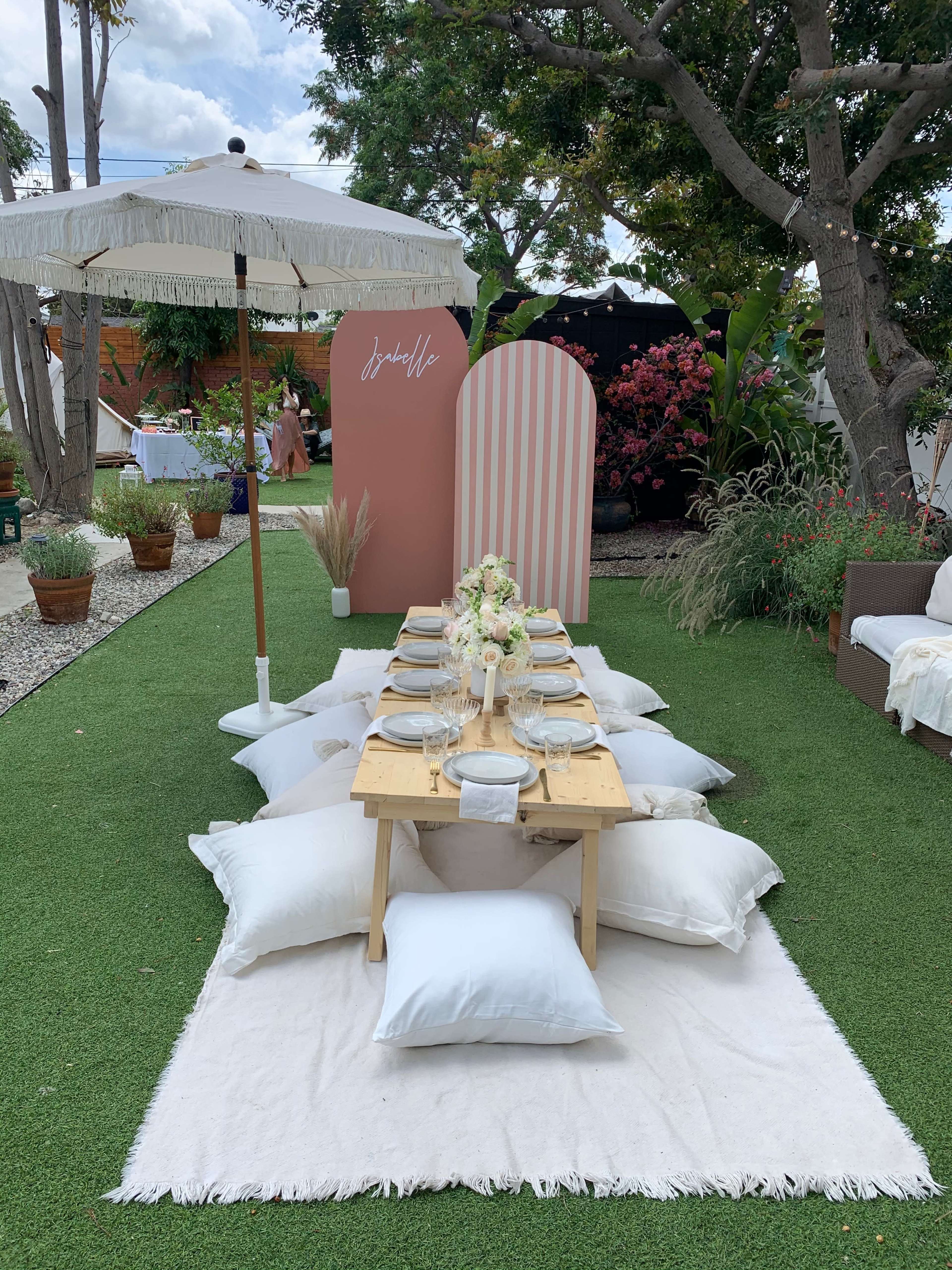 A low table set with plates and cutlery is placed on a white rug surrounded by pillows, with a decorative backdrop and an umbrella in an outdoor garden setting.