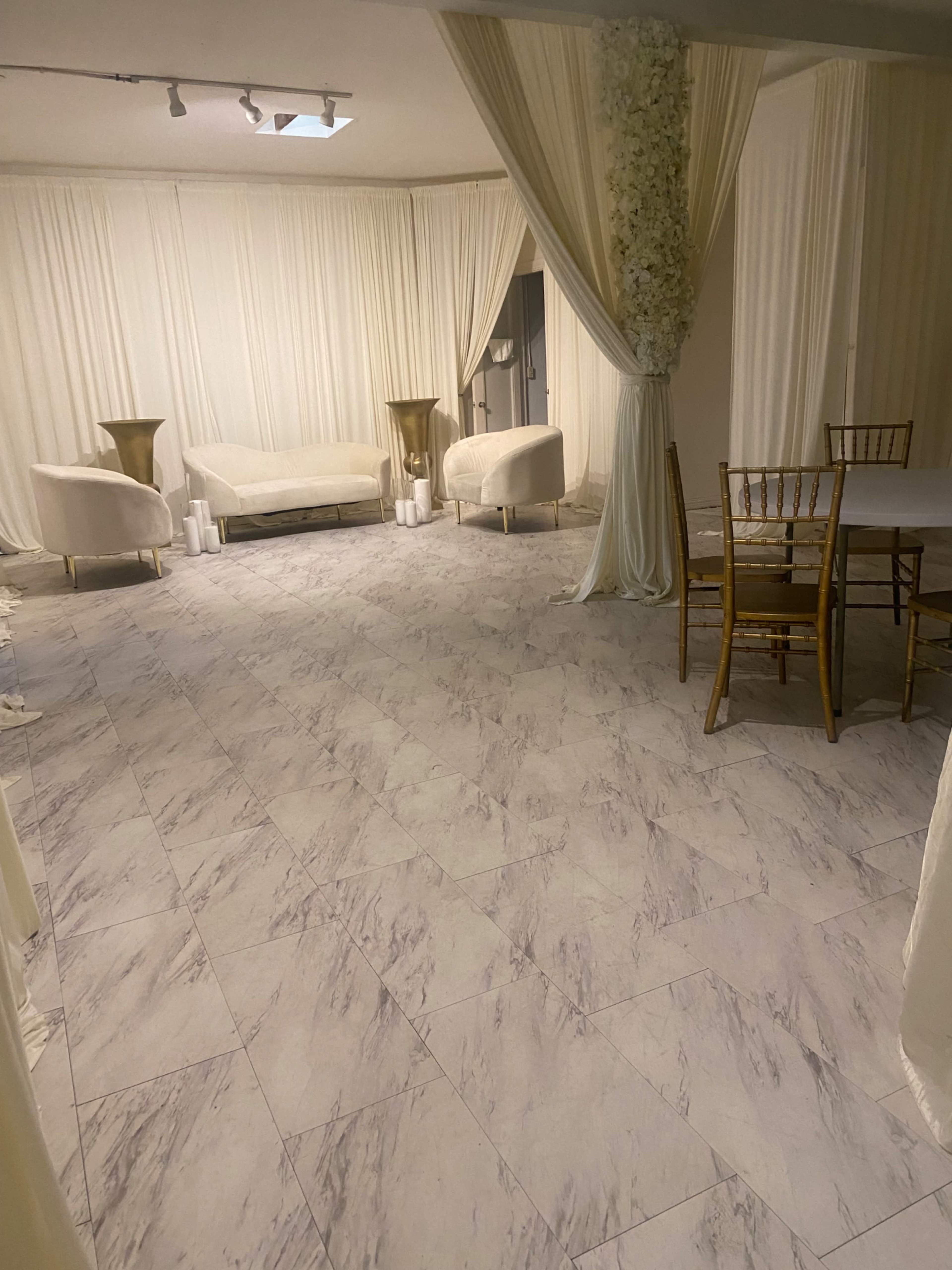 The image shows a room with marble flooring, draped white walls, and two white sofas arranged around a small table, along with gold accents in the decor.