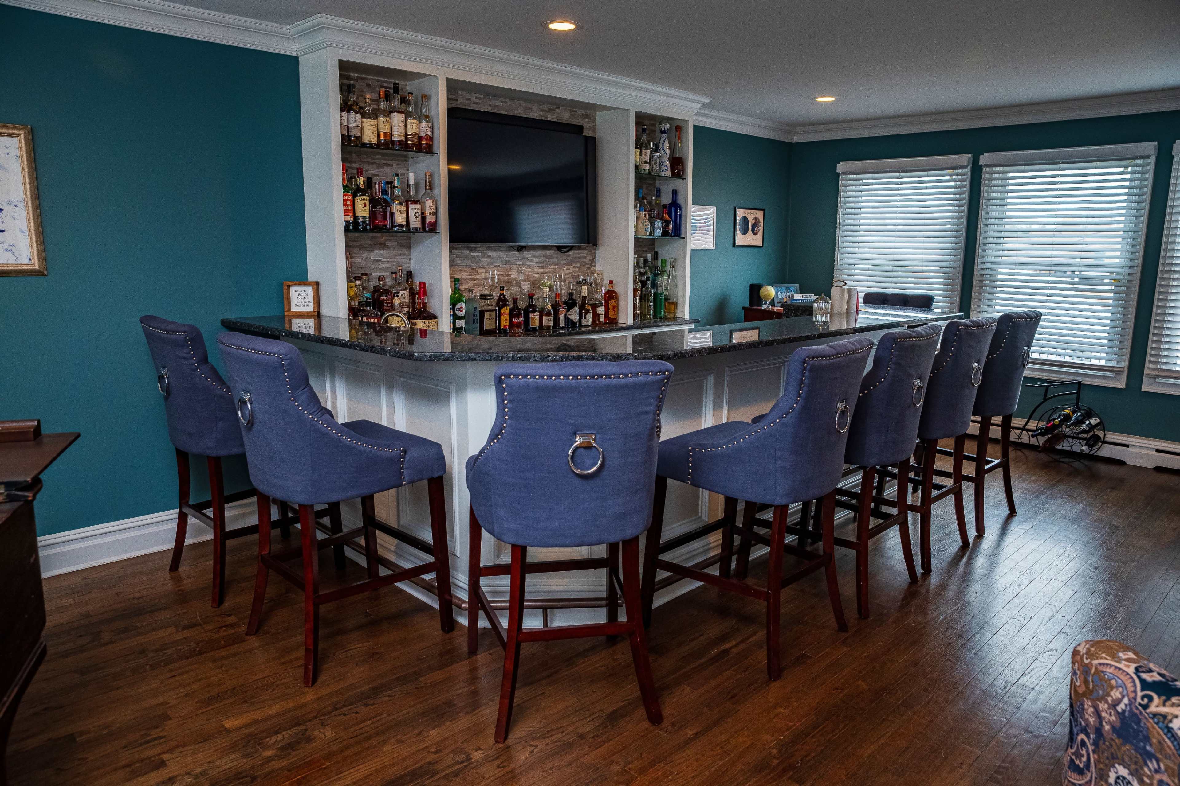 The image shows a home bar area with a counter surrounded by eight upholstered bar stools and a television mounted on the wall, adorned with various bottles and liquor on shelves.