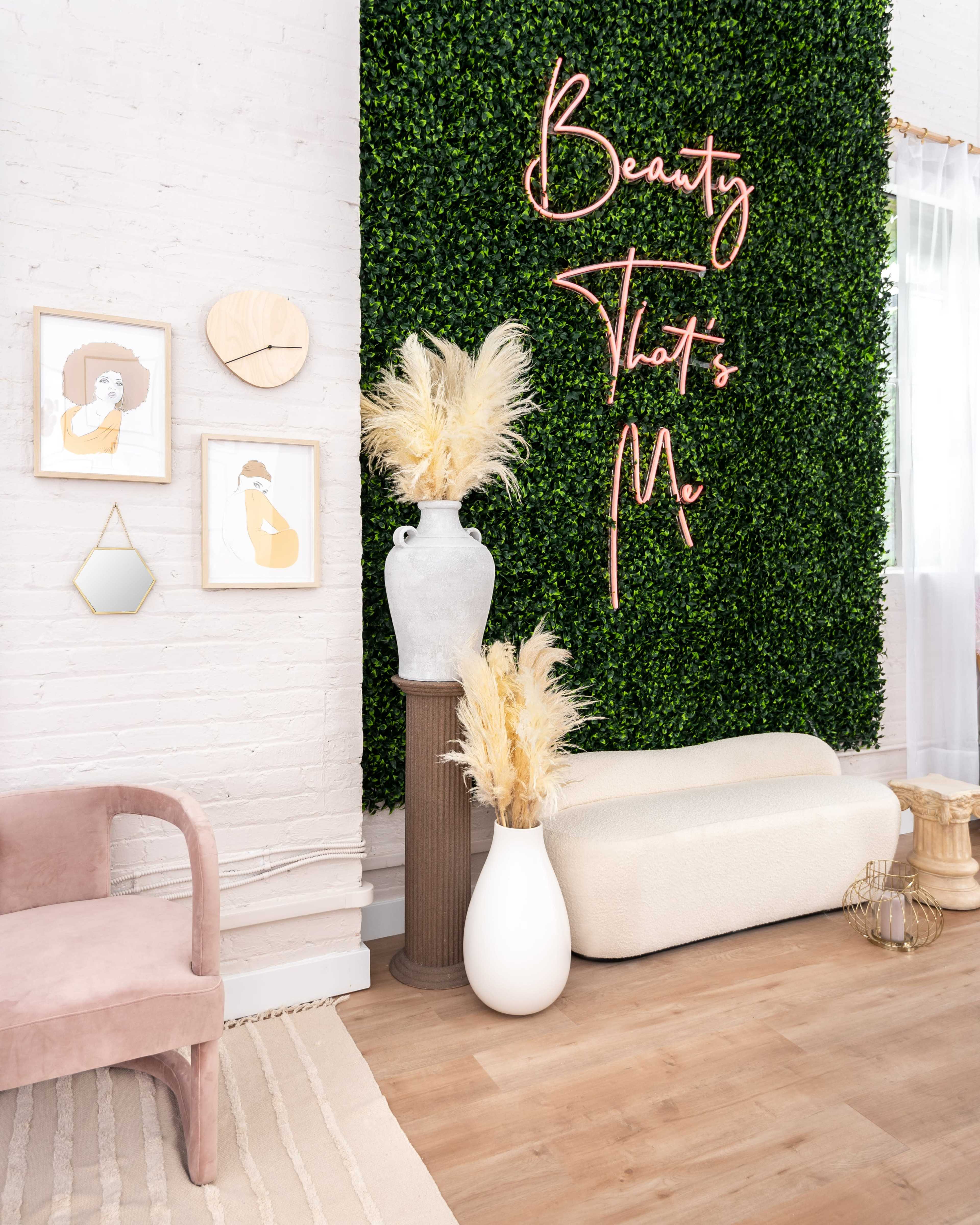 A stylish interior with a green hedge wall, framed art pieces, a white vase, and decorative elements on a wooden floor.