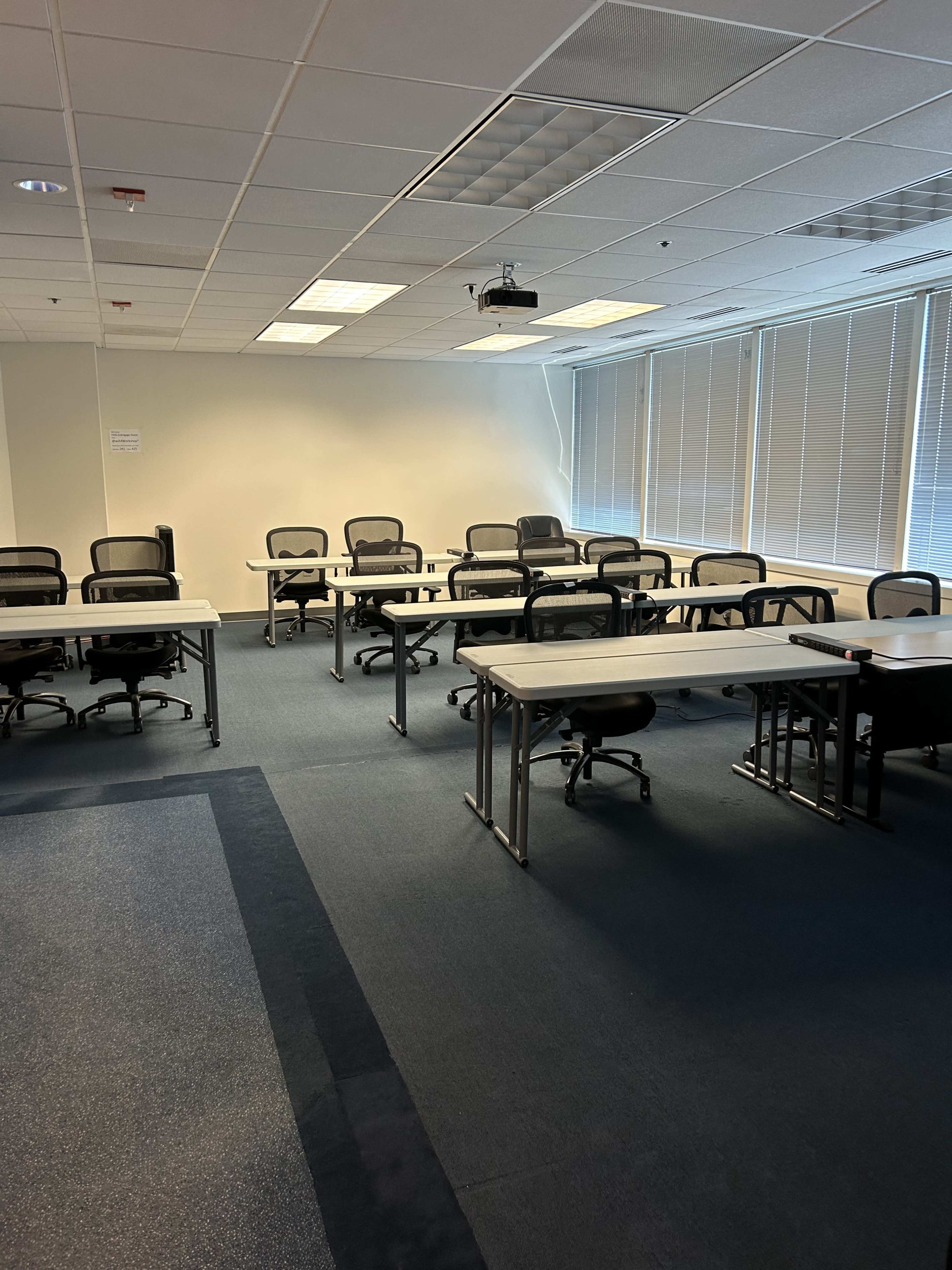 A sparsely furnished meeting room with several tables and chairs arranged in a classroom layout, featuring windows along one side.
