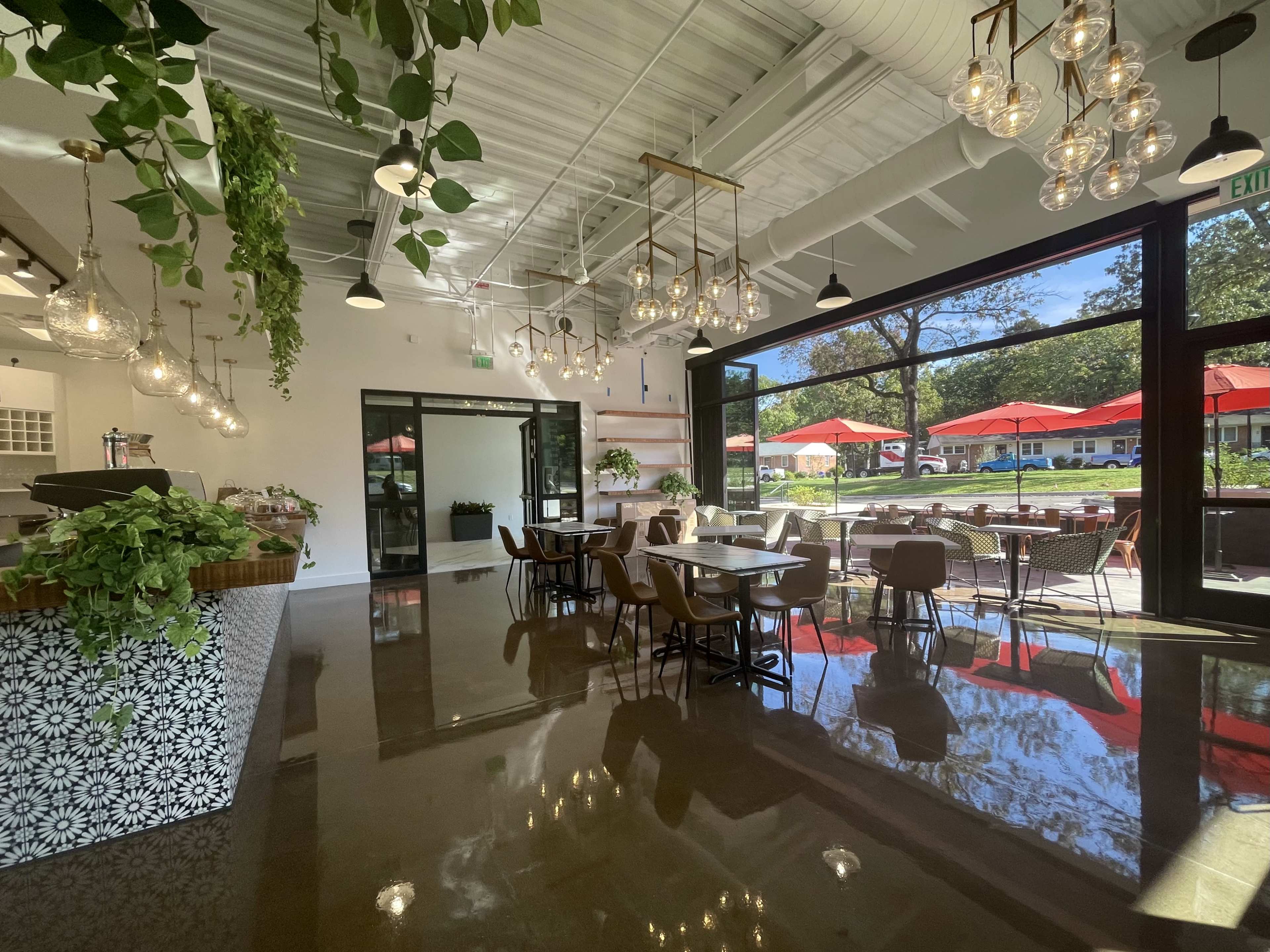 A modern cafe interior with hanging light fixtures, greenery, and a glass wall leading to an outdoor seating area covered by red umbrellas.