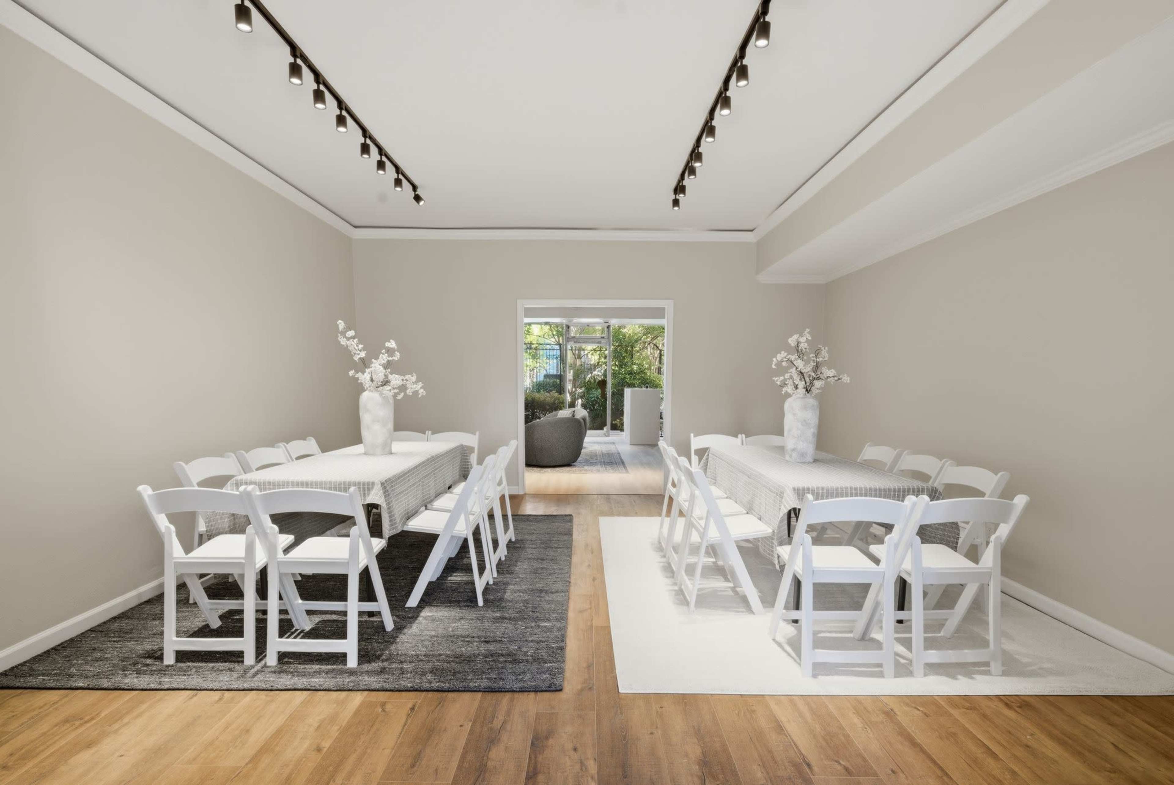The image shows a bright, modern room with two long tables lined with white chairs, surrounded by neutral-colored rugs and large windows that lead to an outdoor area.