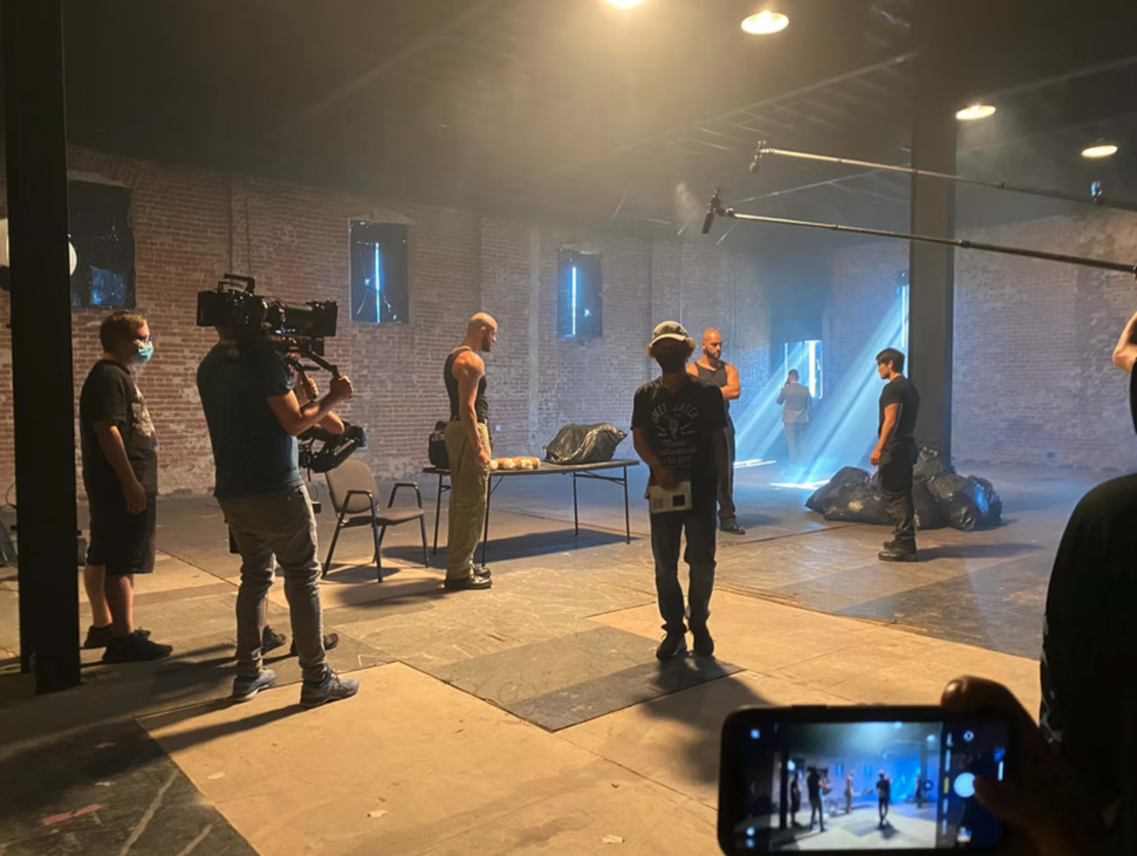 A film crew is capturing a scene in an industrial setting with actors positioned around a table and a camera operator filming from the side.