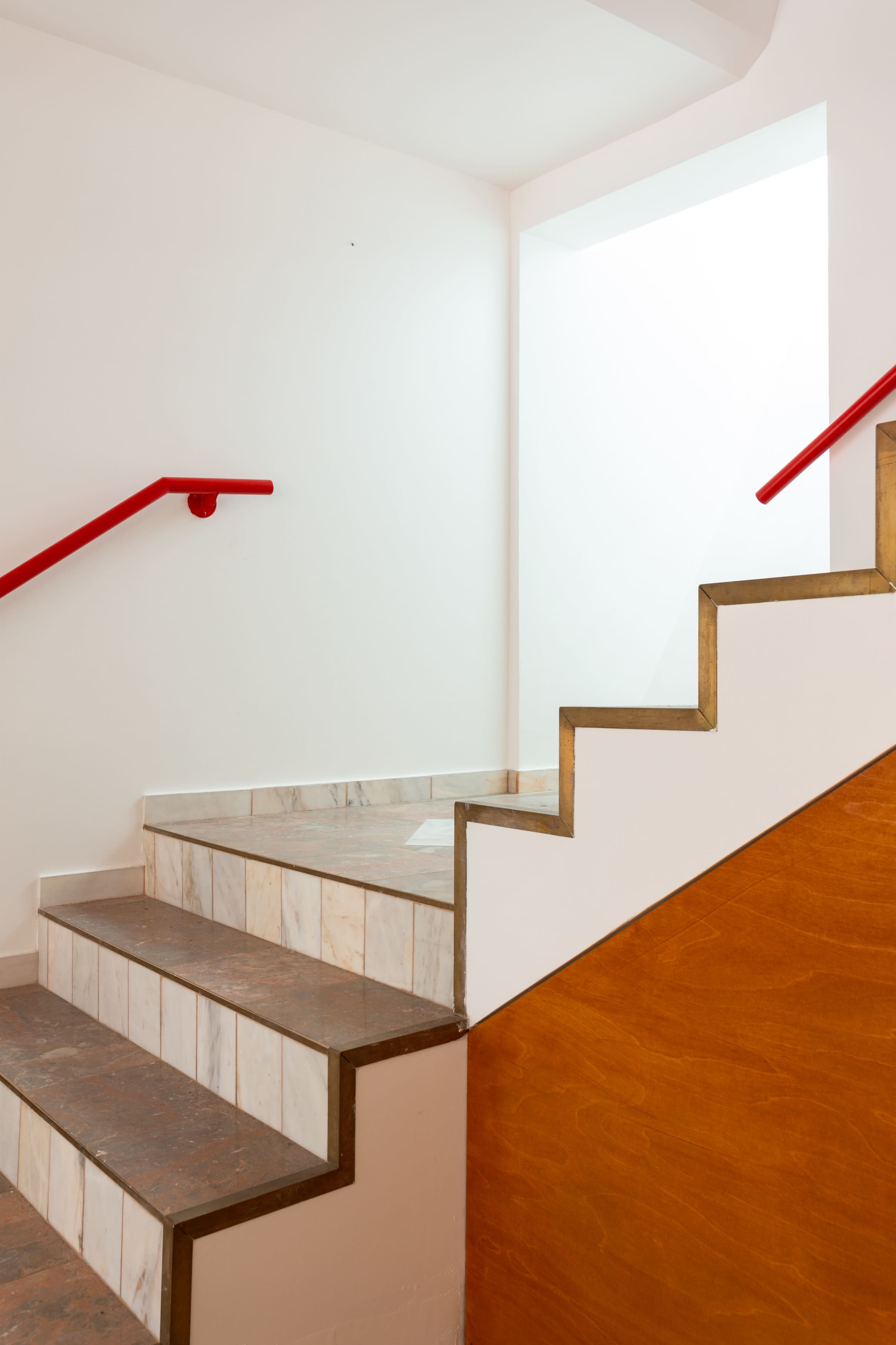 The image shows a stairway with a white wall, wooden accents, and red handrails leading to an upper landing.