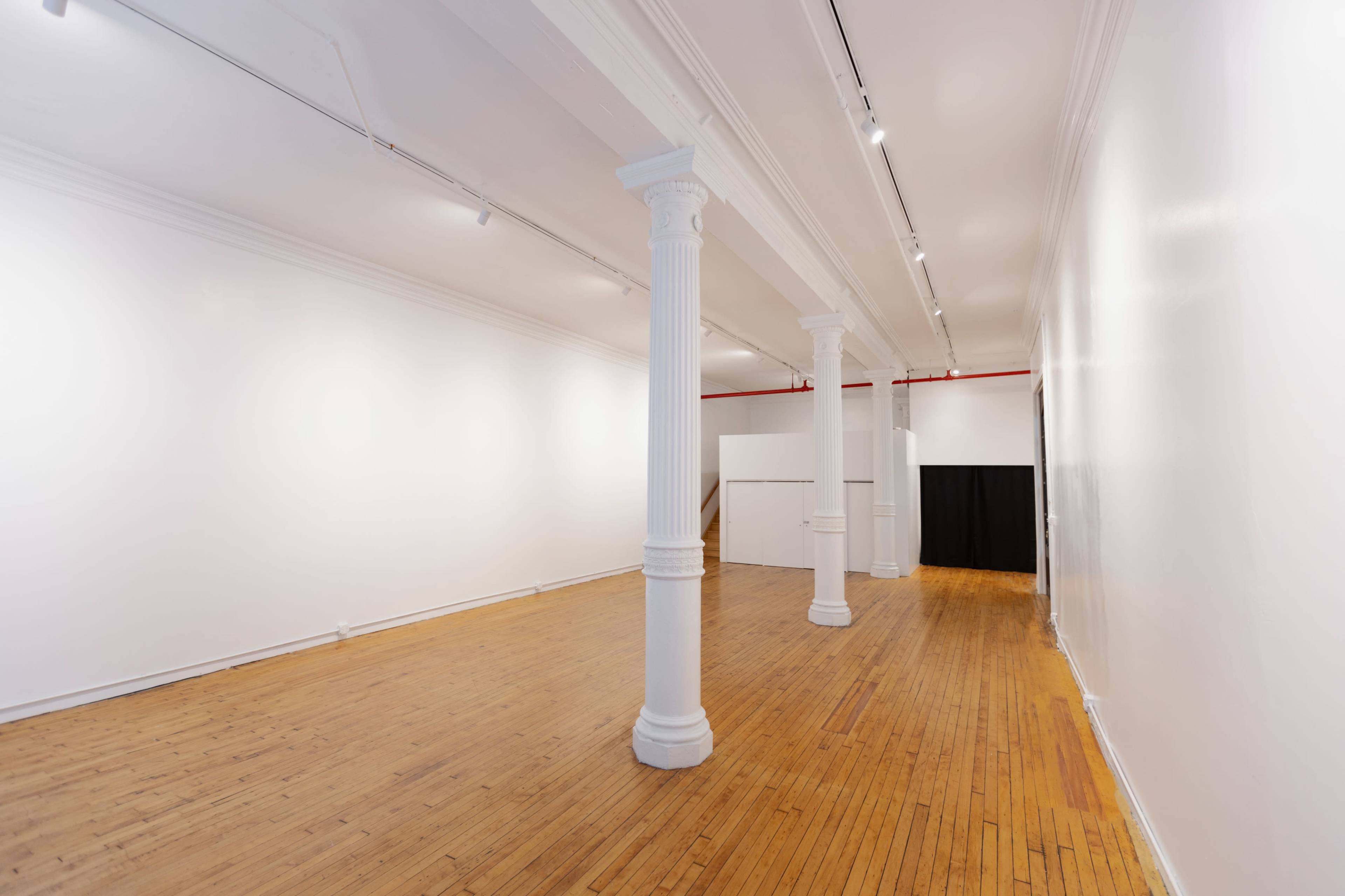 The image shows a spacious art gallery with wooden flooring, white walls, and several columns supporting the ceiling.