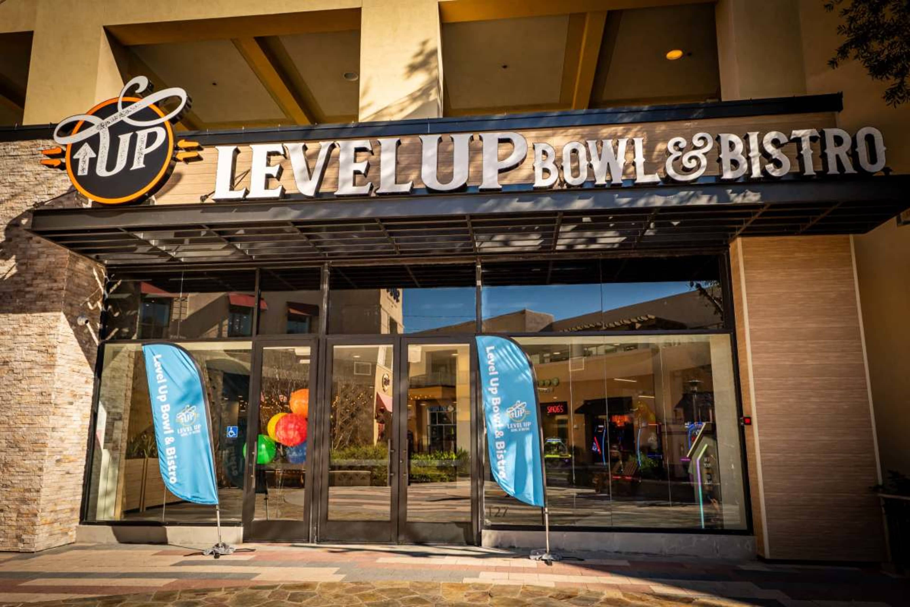 The entrance of Level Up Bowl & Bistro features a large sign with the establishment's name and two blue banners beside the doors.