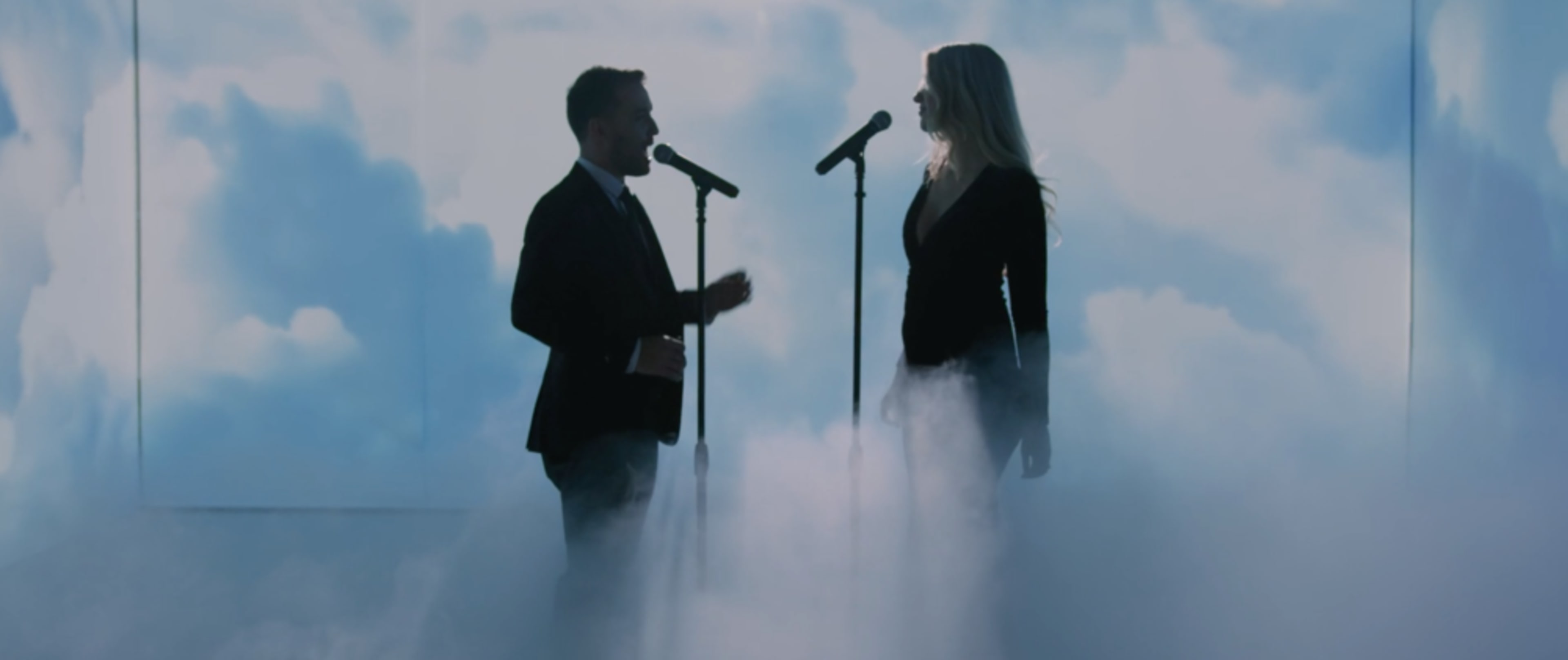 Two singers perform on stage in front of microphones, surrounded by a backdrop of clouds and fog.