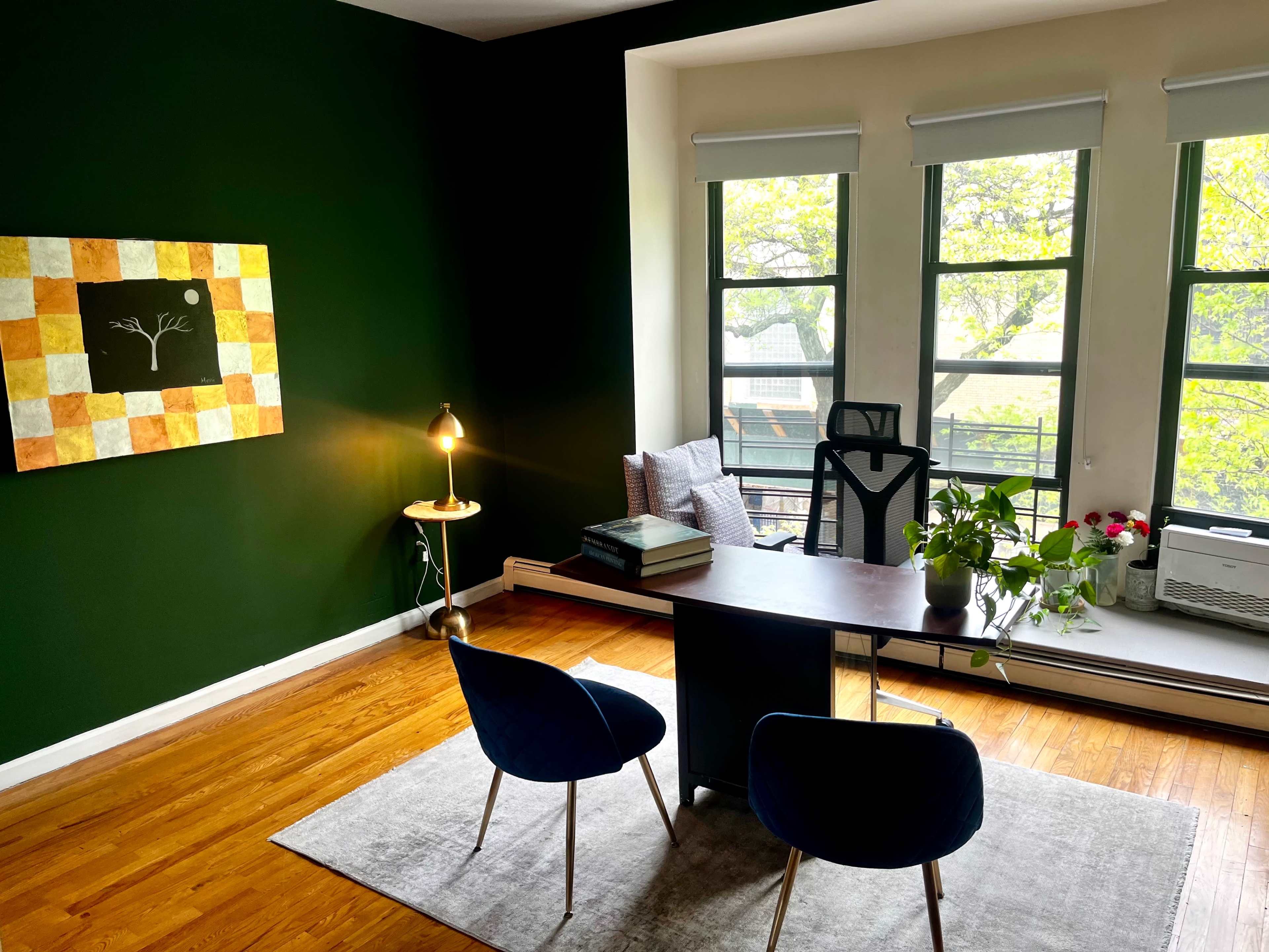 A modern office space features a dark green wall, a wooden desk with books, two blue chairs, and large windows allowing natural light in.