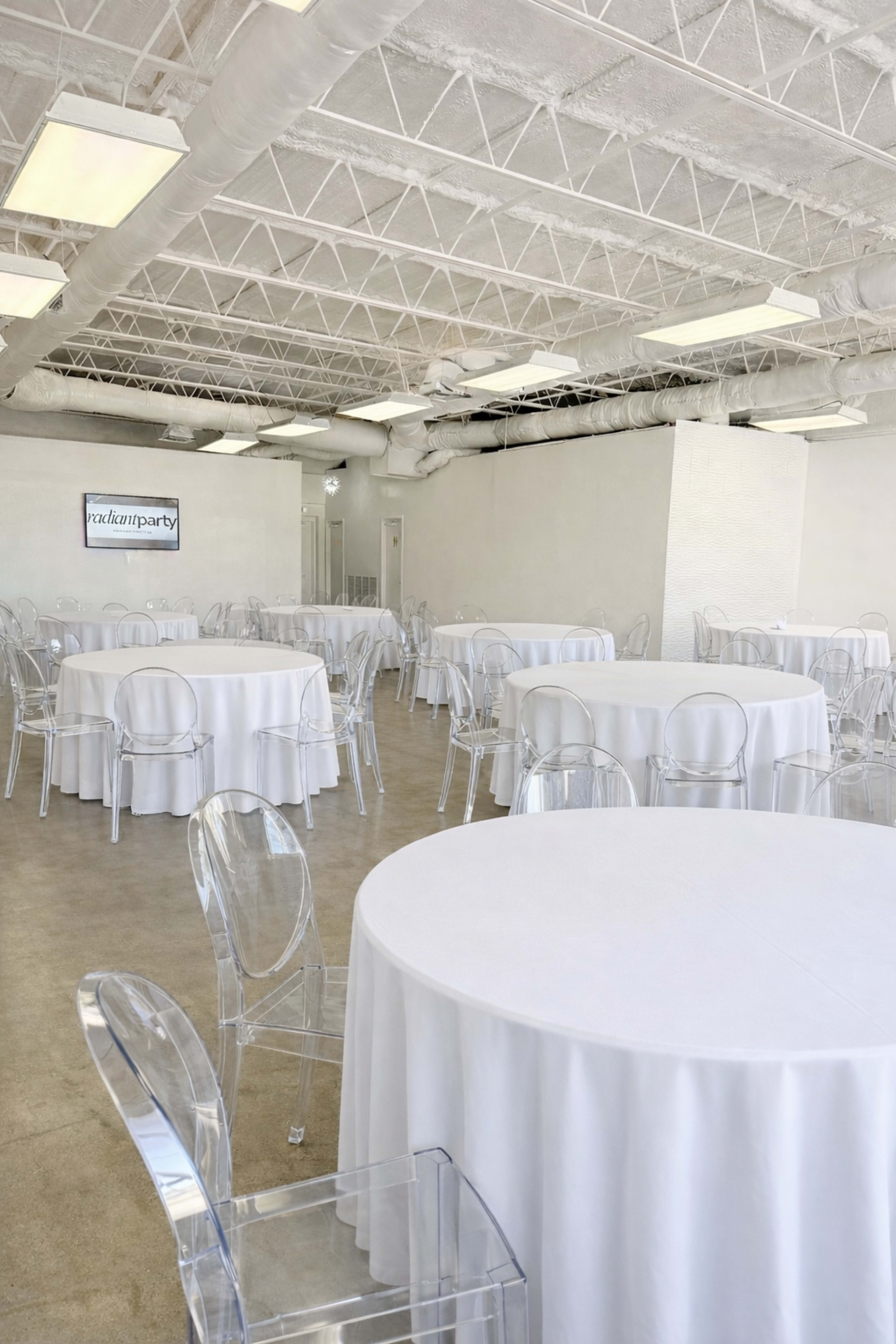 The image shows a spacious, brightly lit event room with several round tables covered in white tablecloths and transparent chairs arranged throughout.