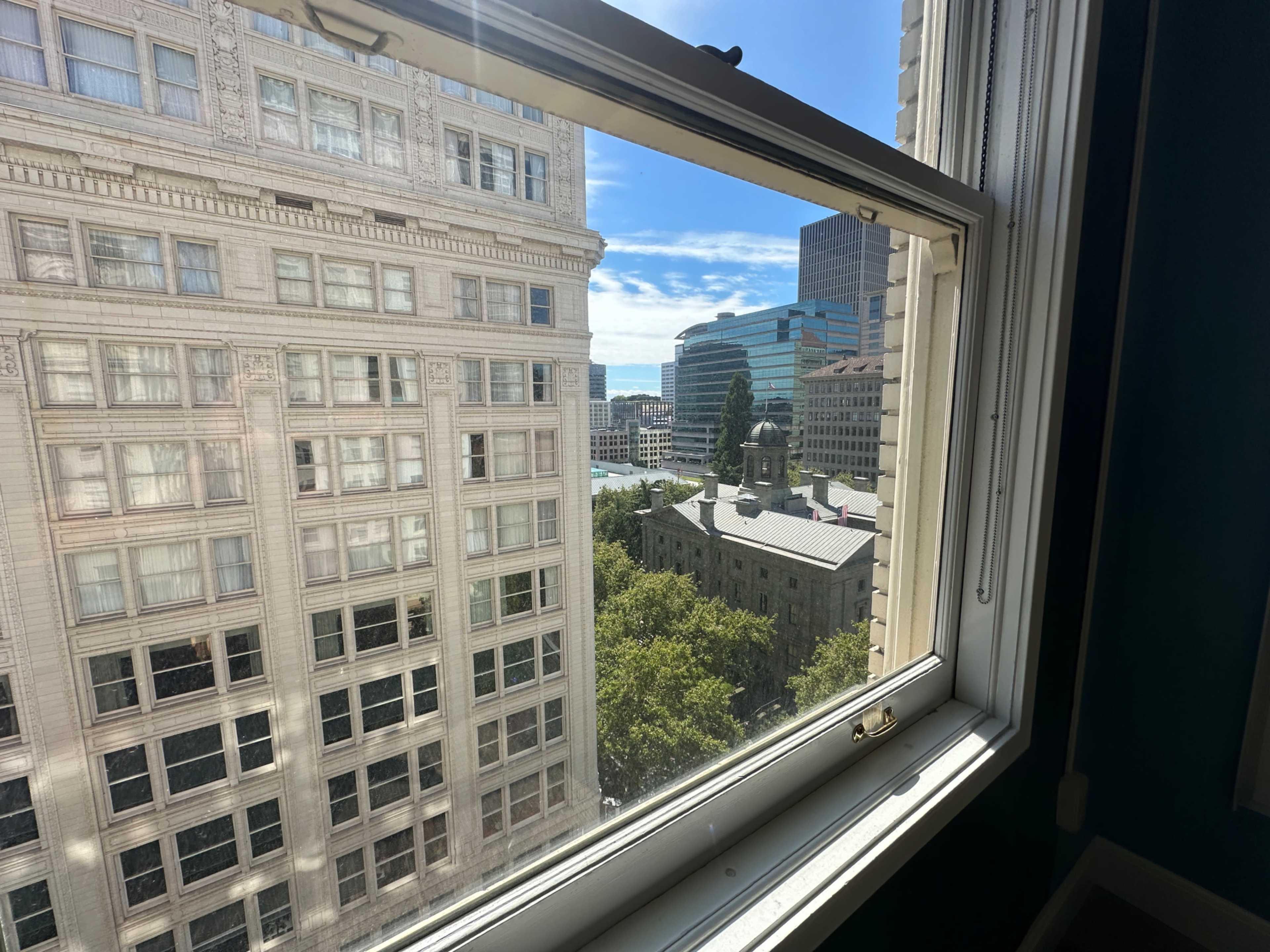 The image shows a view from a building window, capturing neighboring buildings and a park area under a clear blue sky.
