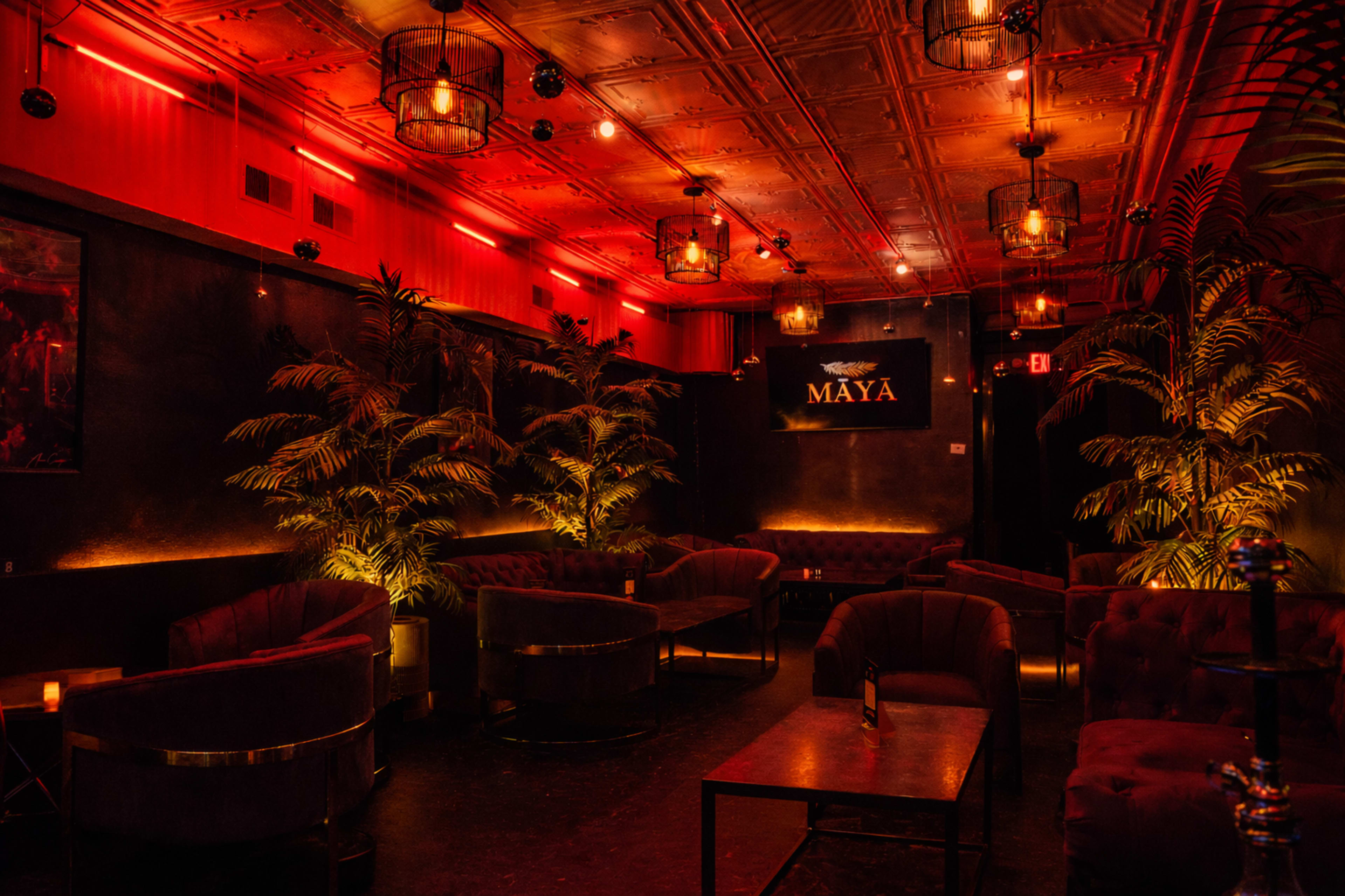 The image depicts a dimly lit lounge with plush seating, illuminated by red light and featuring tall potted plants.