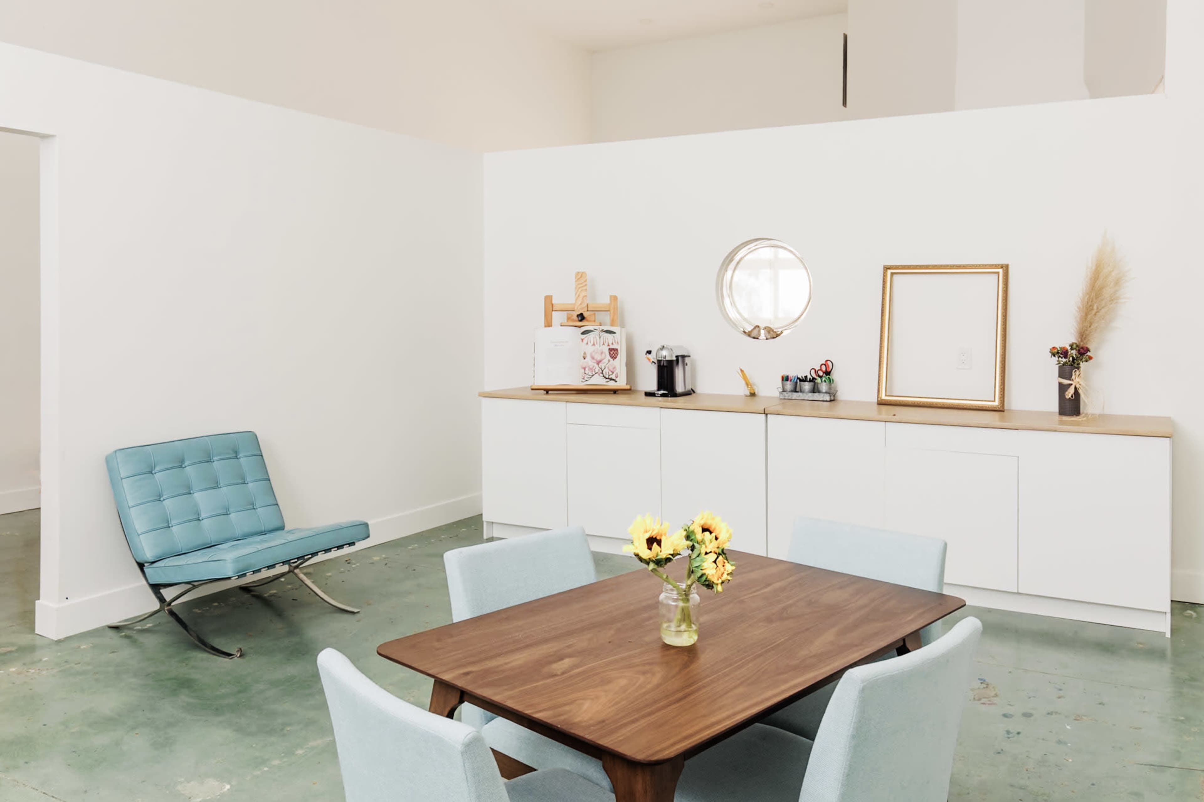 A modern dining area features a wooden table surrounded by light blue chairs, with a minimalist sideboard and a decorative mirror on a white wall.