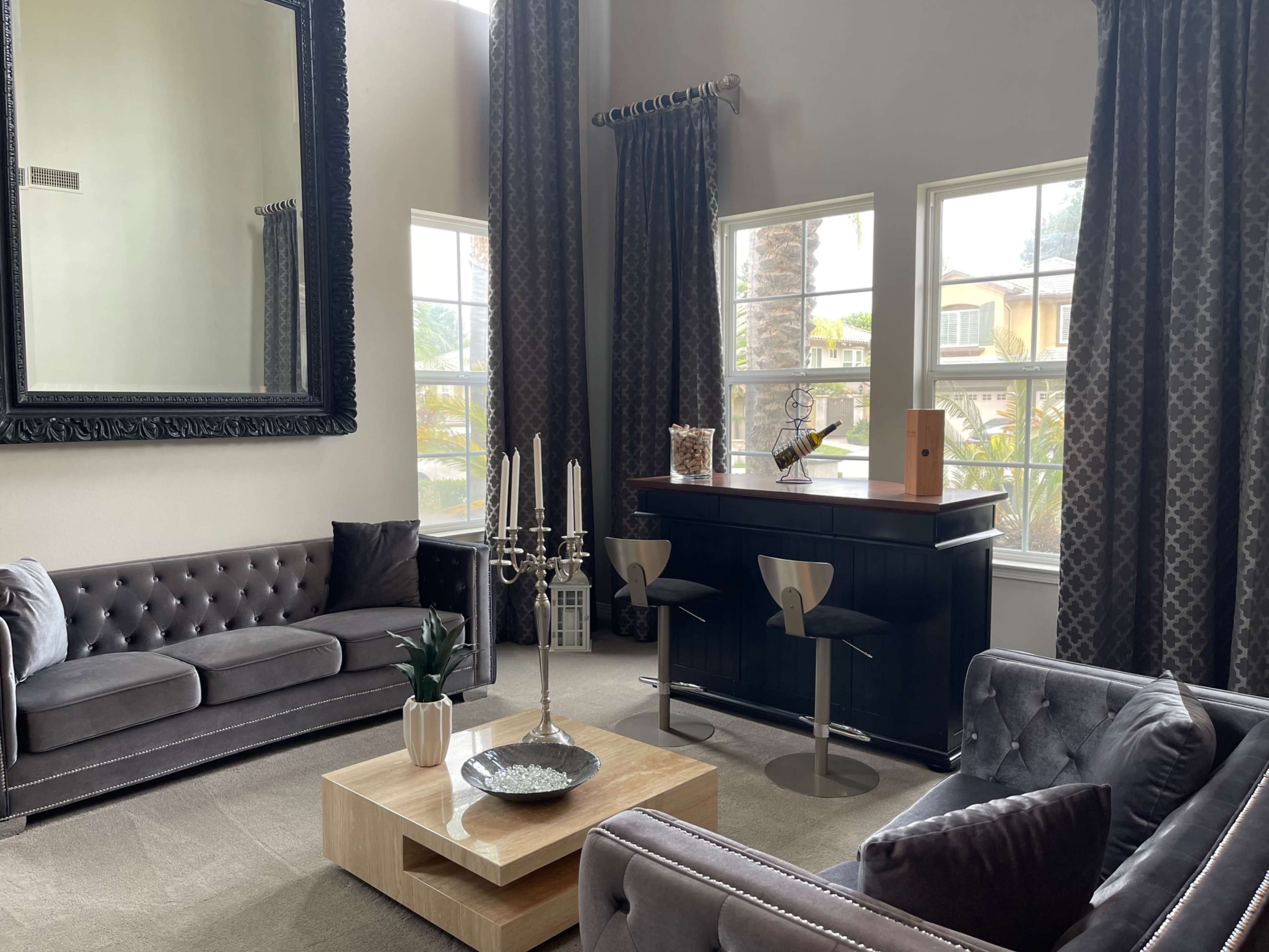 The scene features a modern living room with dark upholstered sofas, a wooden coffee table, a bar area, and large windows allowing natural light to enter.