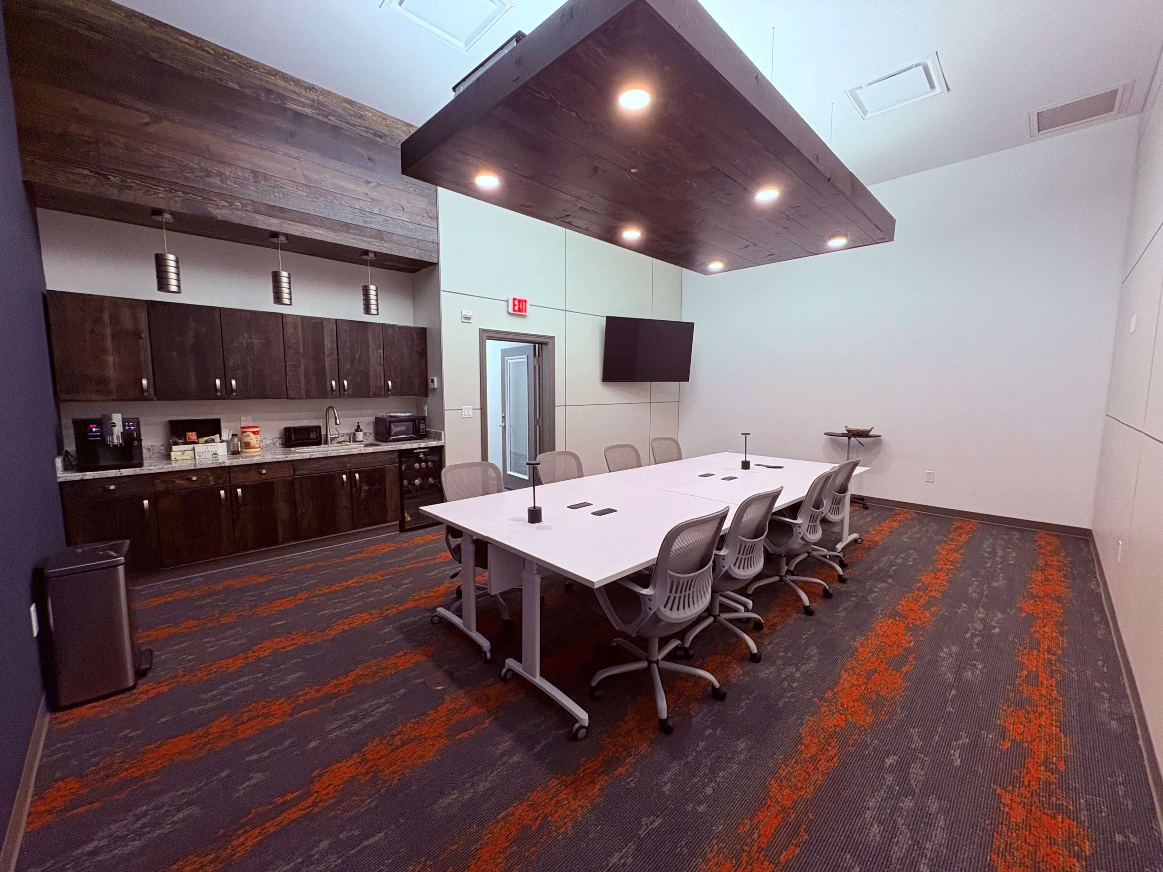 The image shows a modern conference room with a long white table surrounded by chairs, a kitchen area, and a wall-mounted screen.
