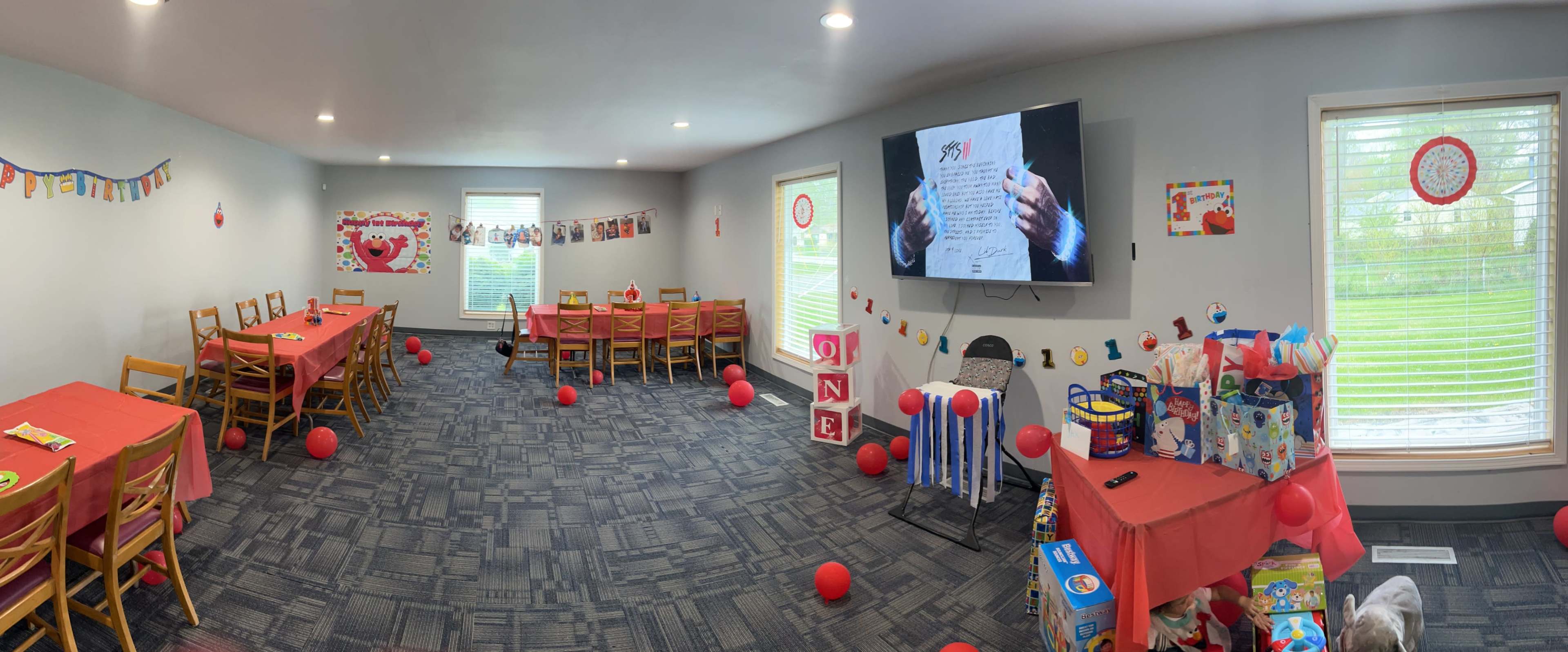 A party room is decorated with red balloons, table settings, and a large screen displaying a document, while a variety of gifts are arranged on a side table.