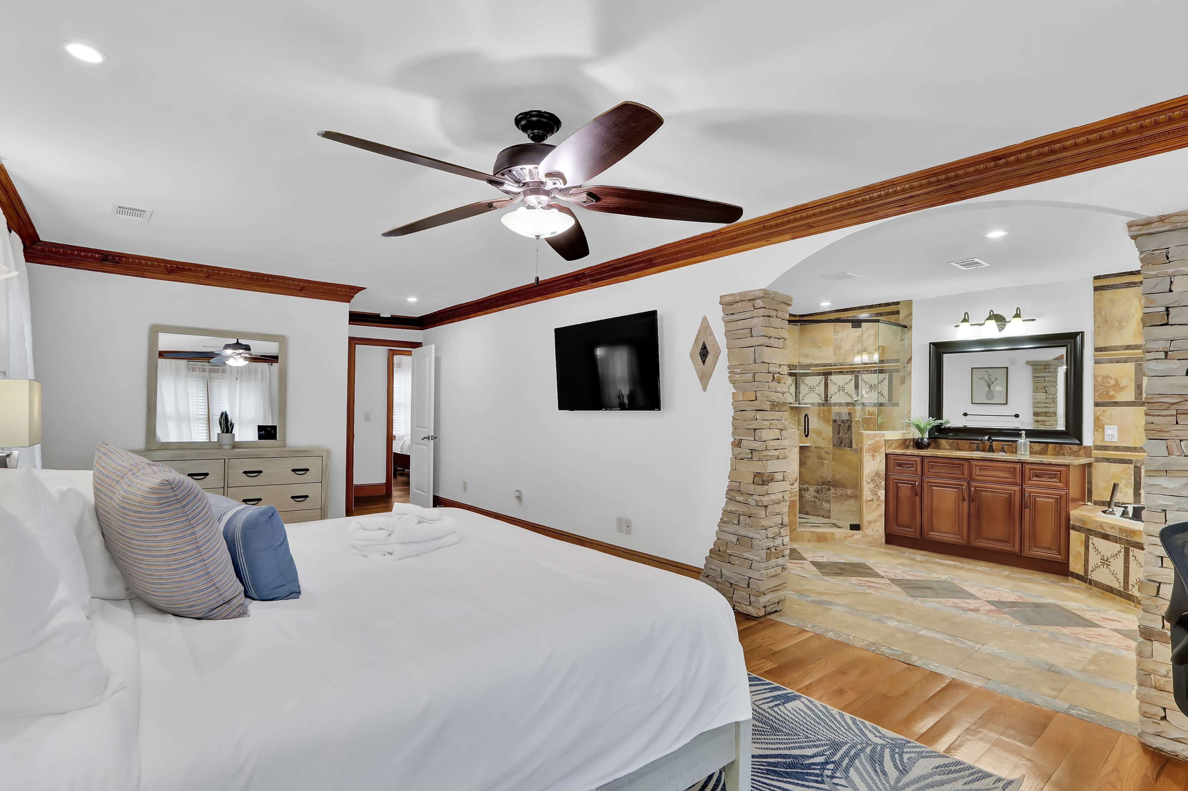 A spacious bedroom with a king-size bed, a ceiling fan, and a view into a bathroom featuring stone accents and wooden cabinetry.