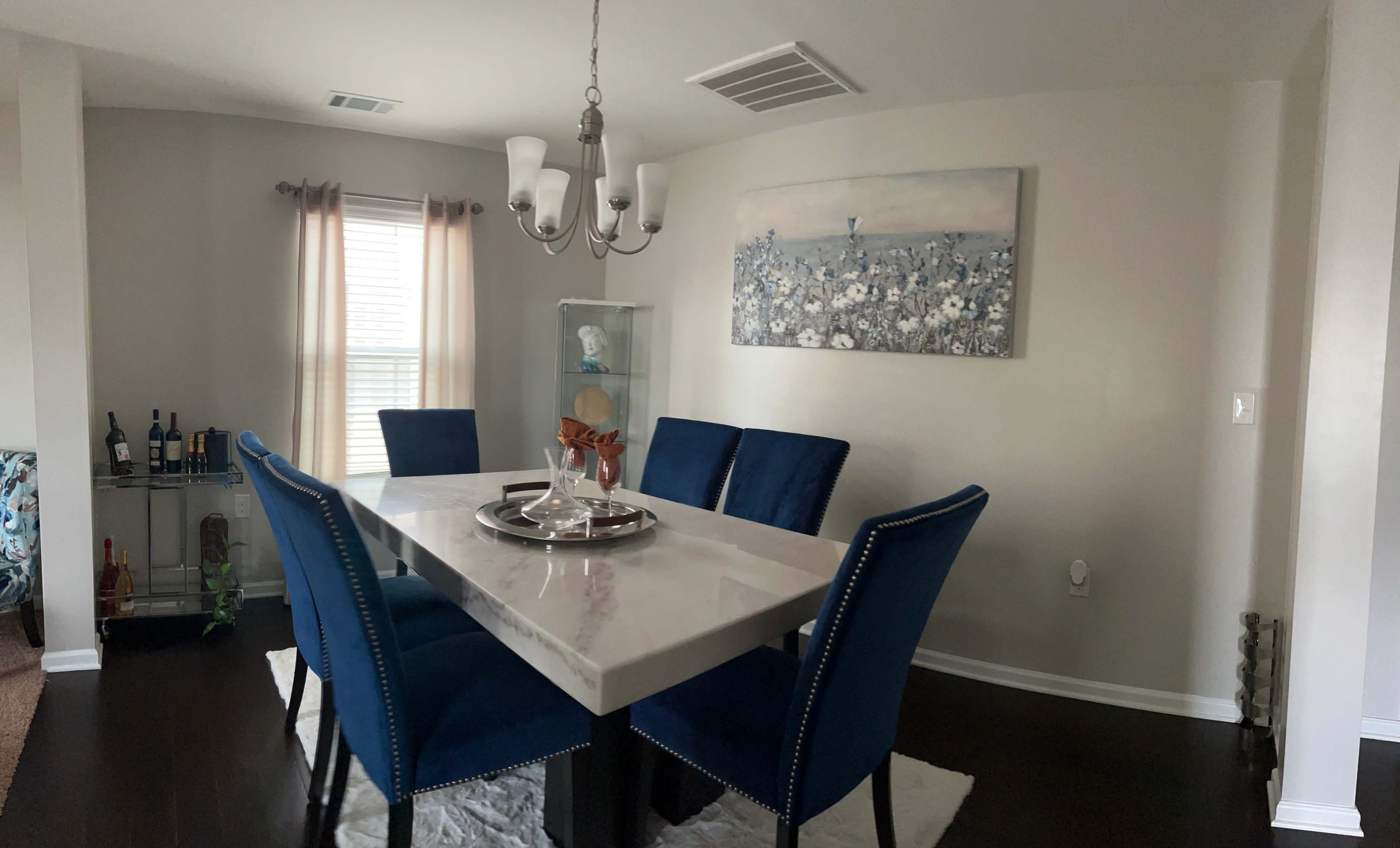A dining area featuring a rectangular table surrounded by blue upholstered chairs, with a chandelier overhead and a floral painting on the wall.