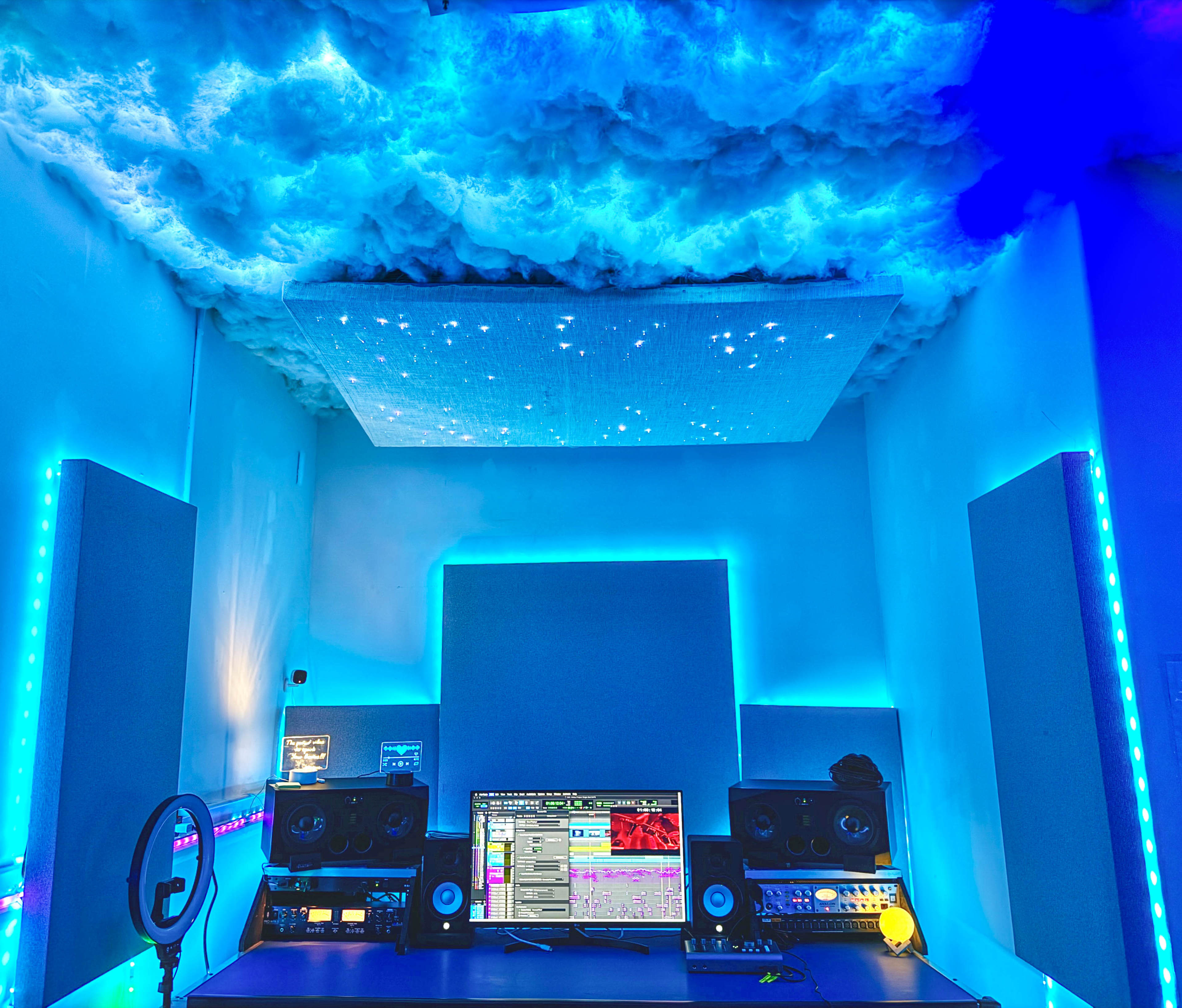 A music production studio with blue lighting, cloud-like ceiling decorations, a central monitor, and sound equipment on a desk.