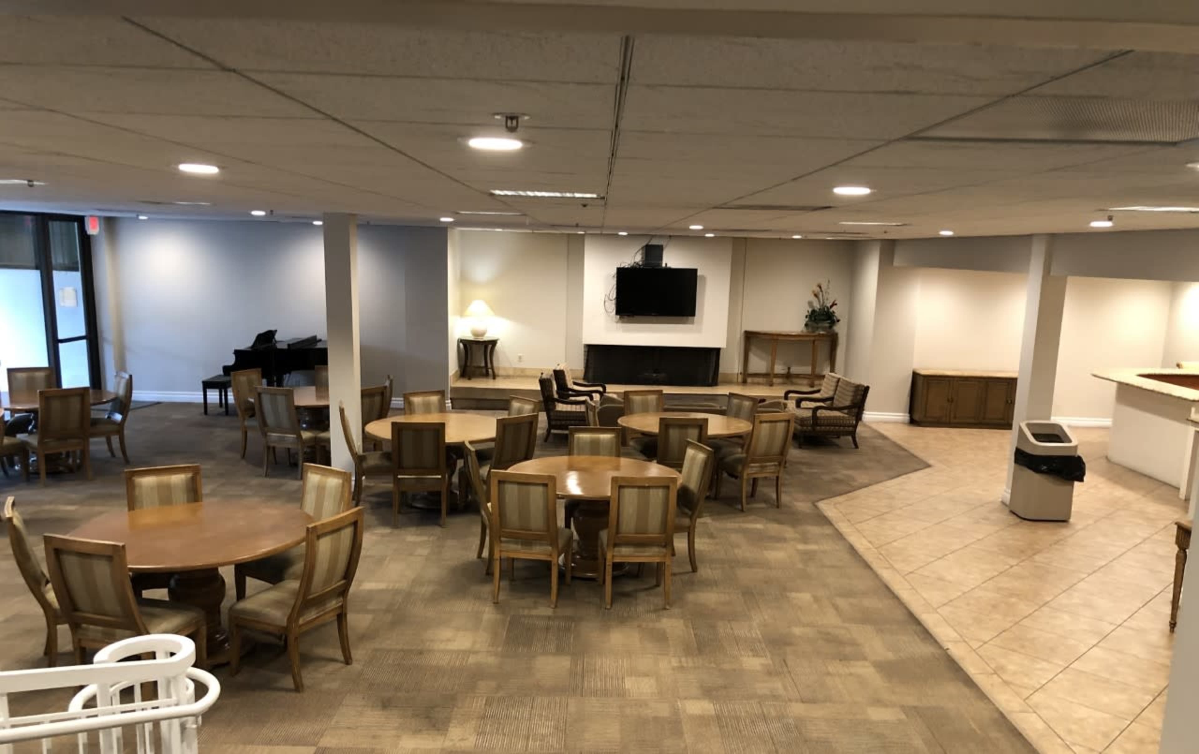 A spacious community room with several tables and chairs, a piano, a television on the wall, and a kitchenette area.
