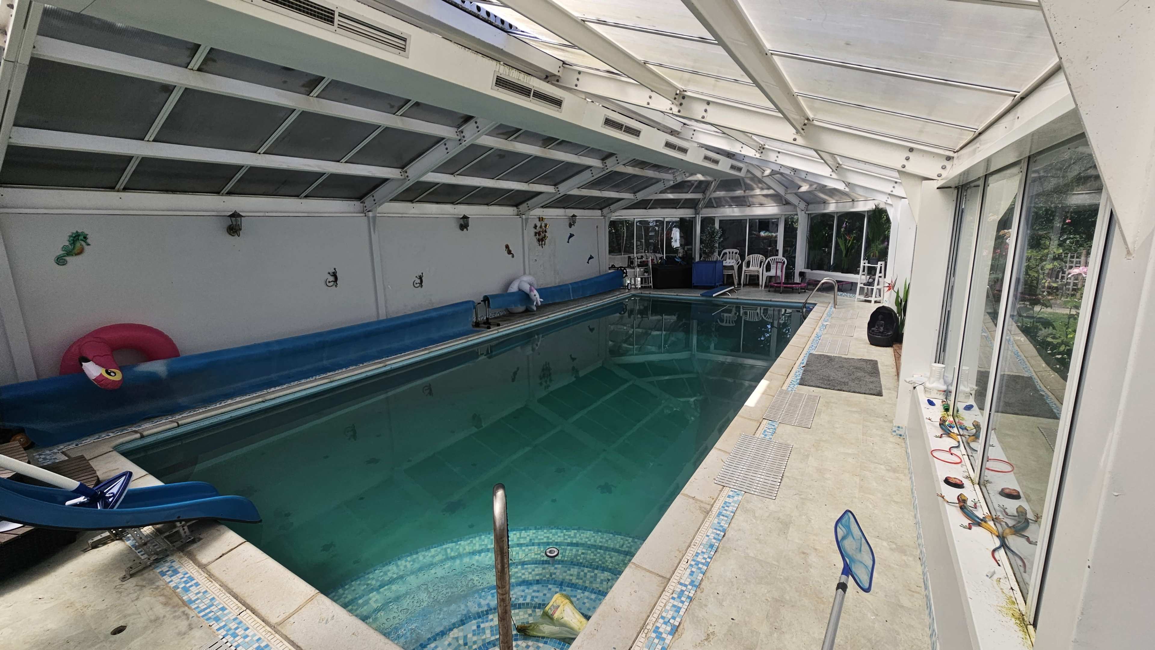 The image shows an indoor swimming pool enclosed by a clear roof, featuring a water slide, lounge chairs, and a landscaped view through large windows.