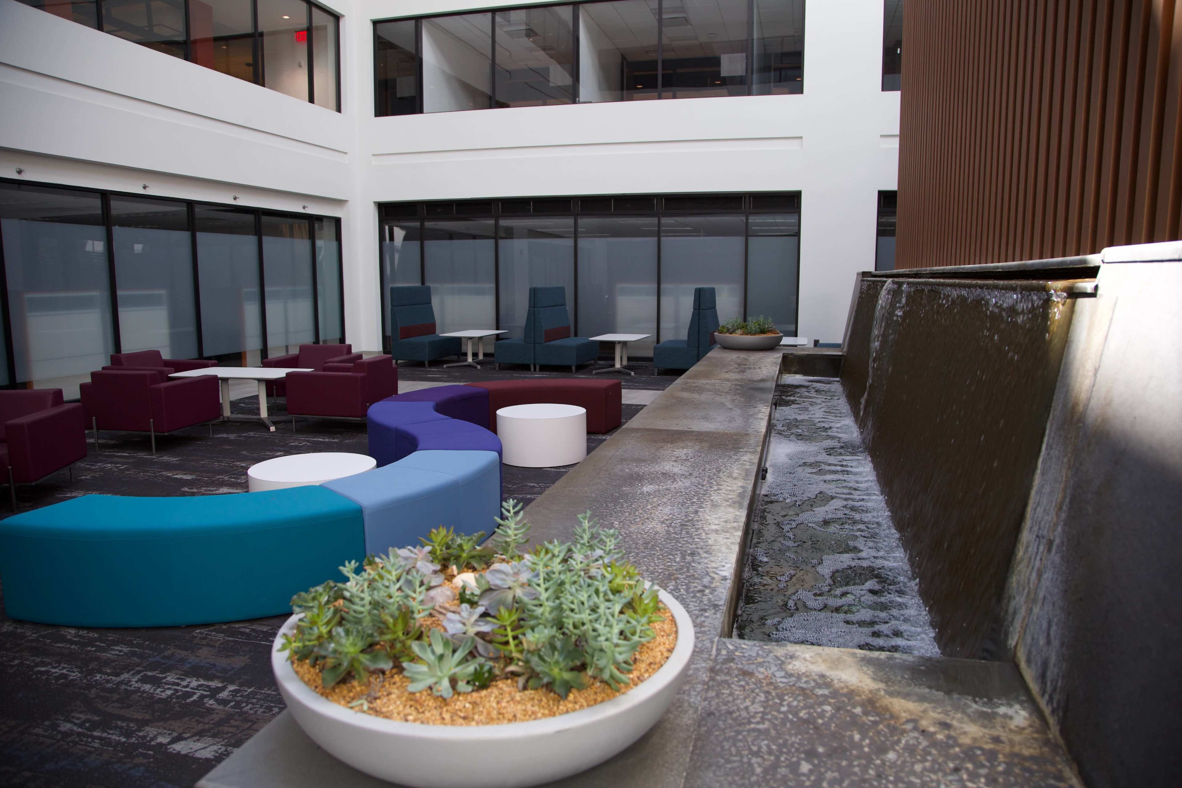 The image shows a modern interior courtyard with seating areas featuring colorful couches and a water feature beside potted plants.