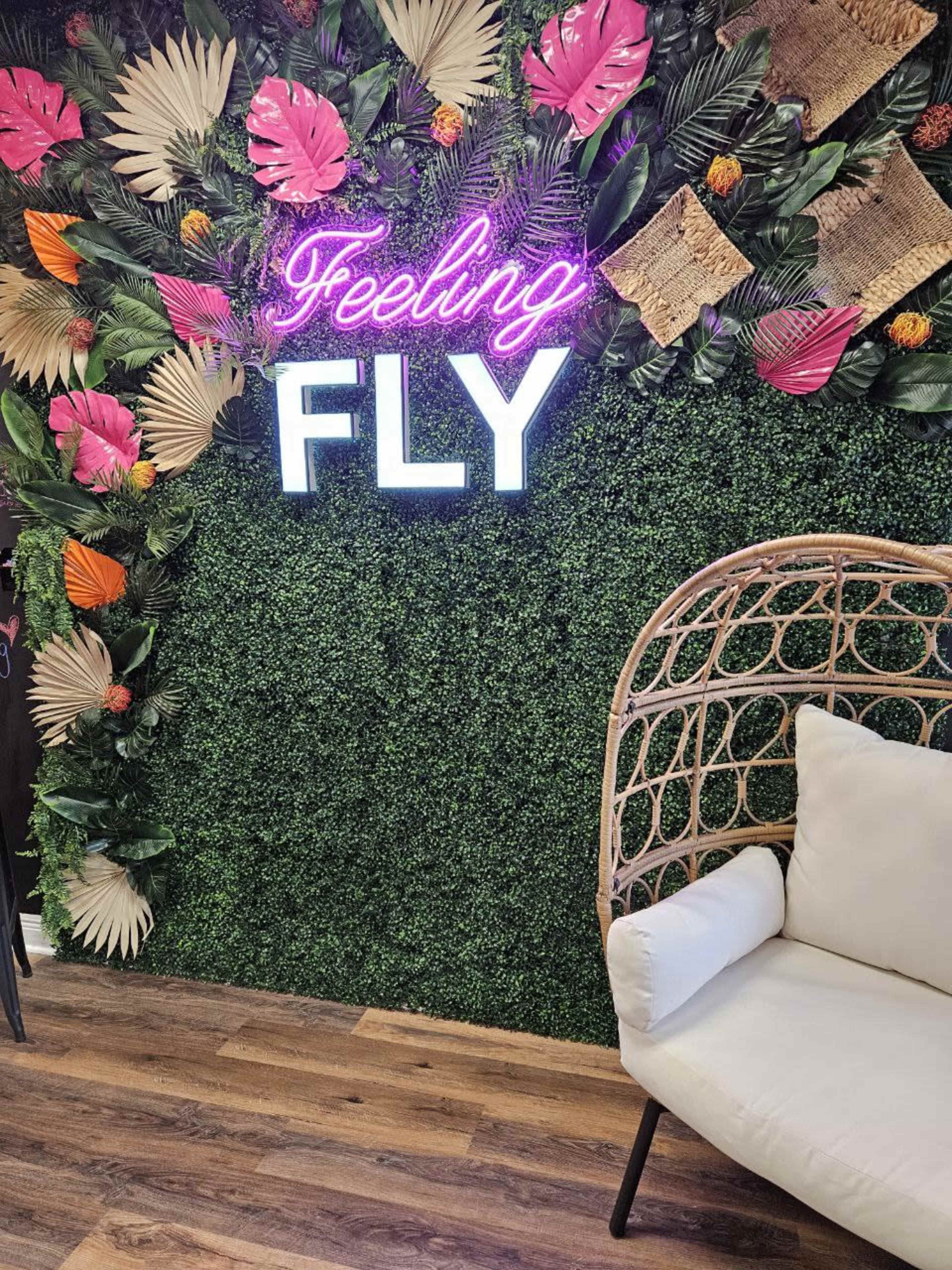 A lush green wall adorned with tropical leaves and flowers, alongside a neon sign that reads "Feeling FLY," and a wicker chair nearby.