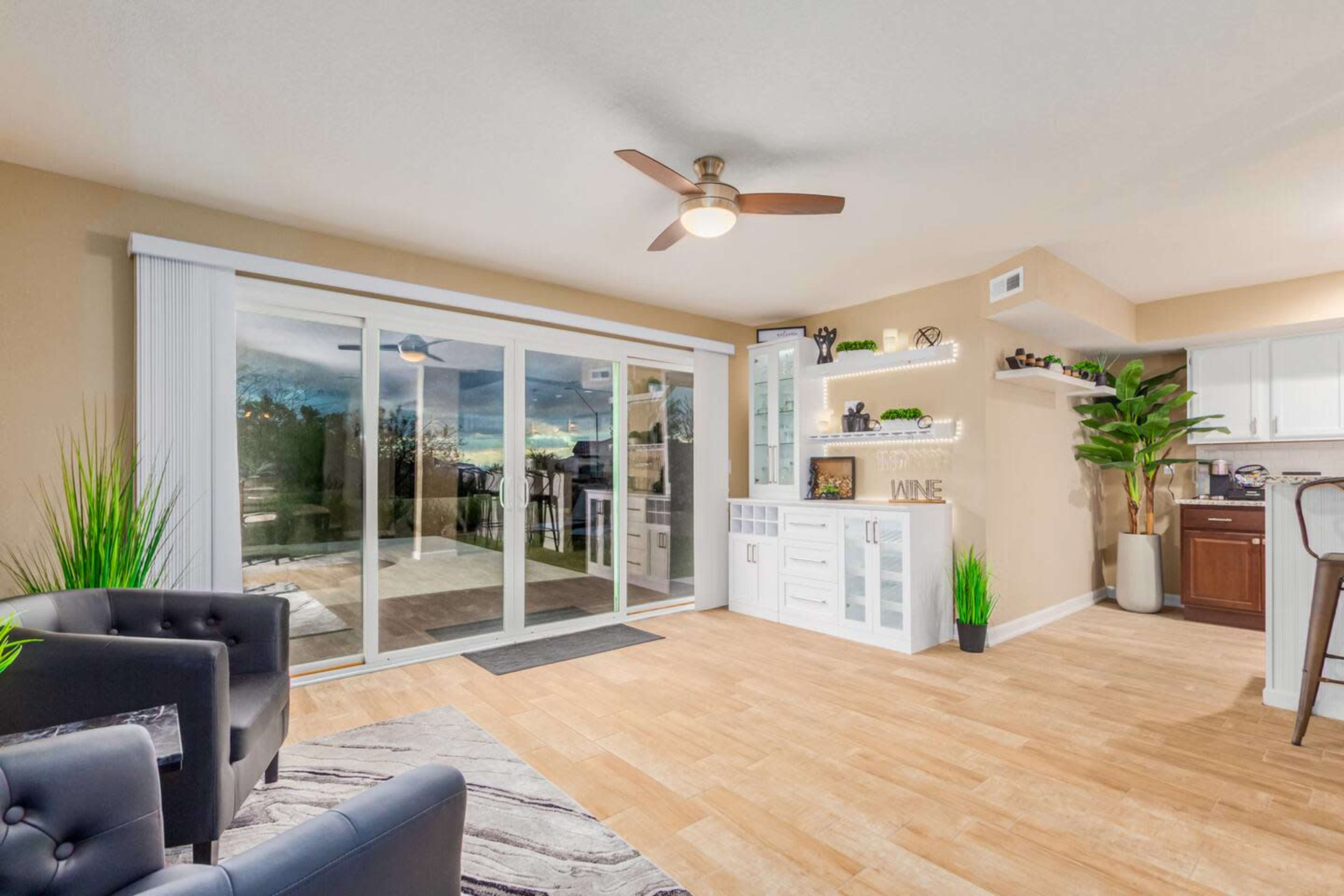 The image shows a modern living room with a sliding glass door that opens to an outdoor space, featuring a kitchenette area, a ceiling fan, and decorative plants.