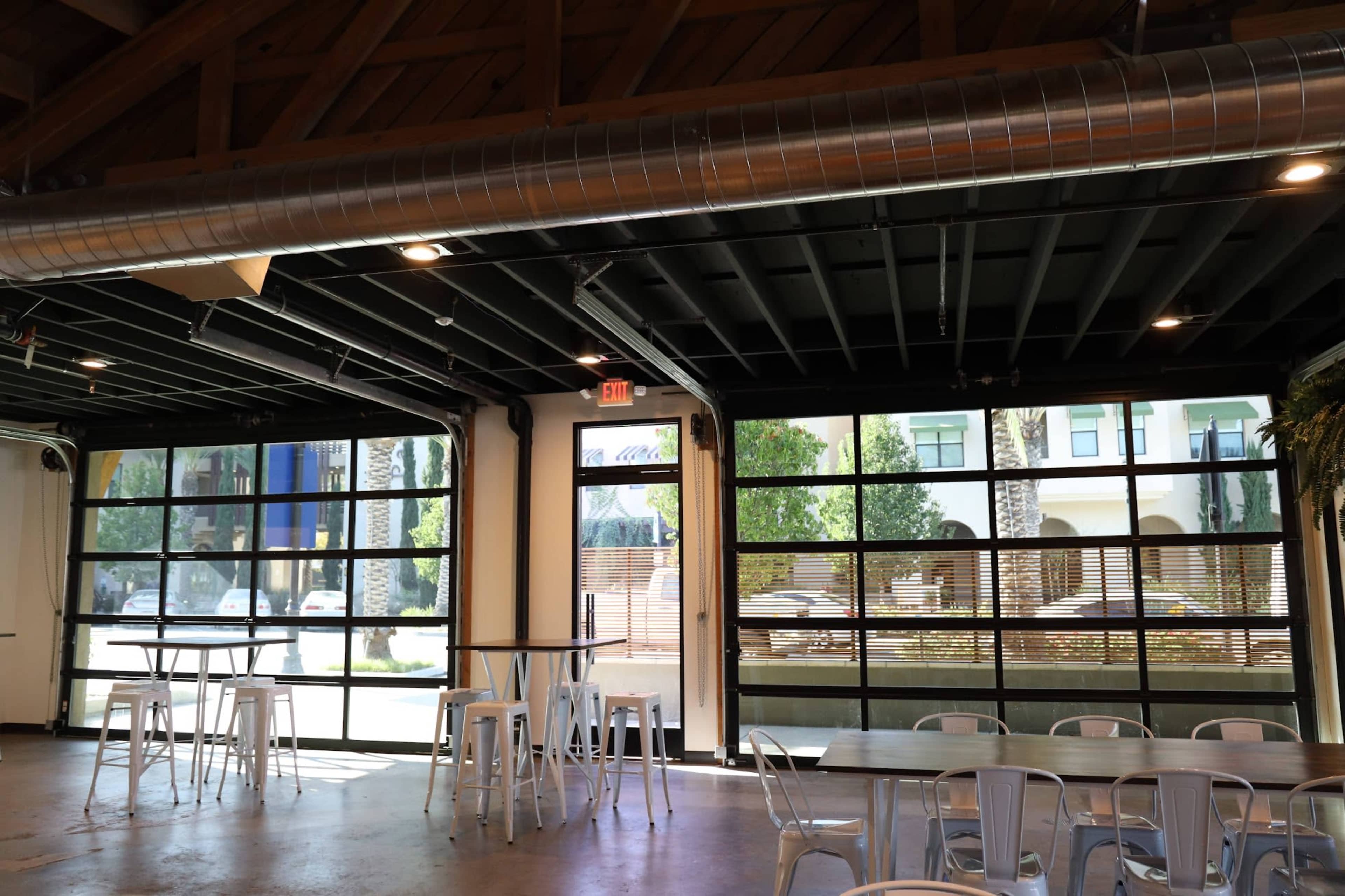 The image shows an interior space with large glass garage doors, modern tables and chairs, and a view of greenery outside.