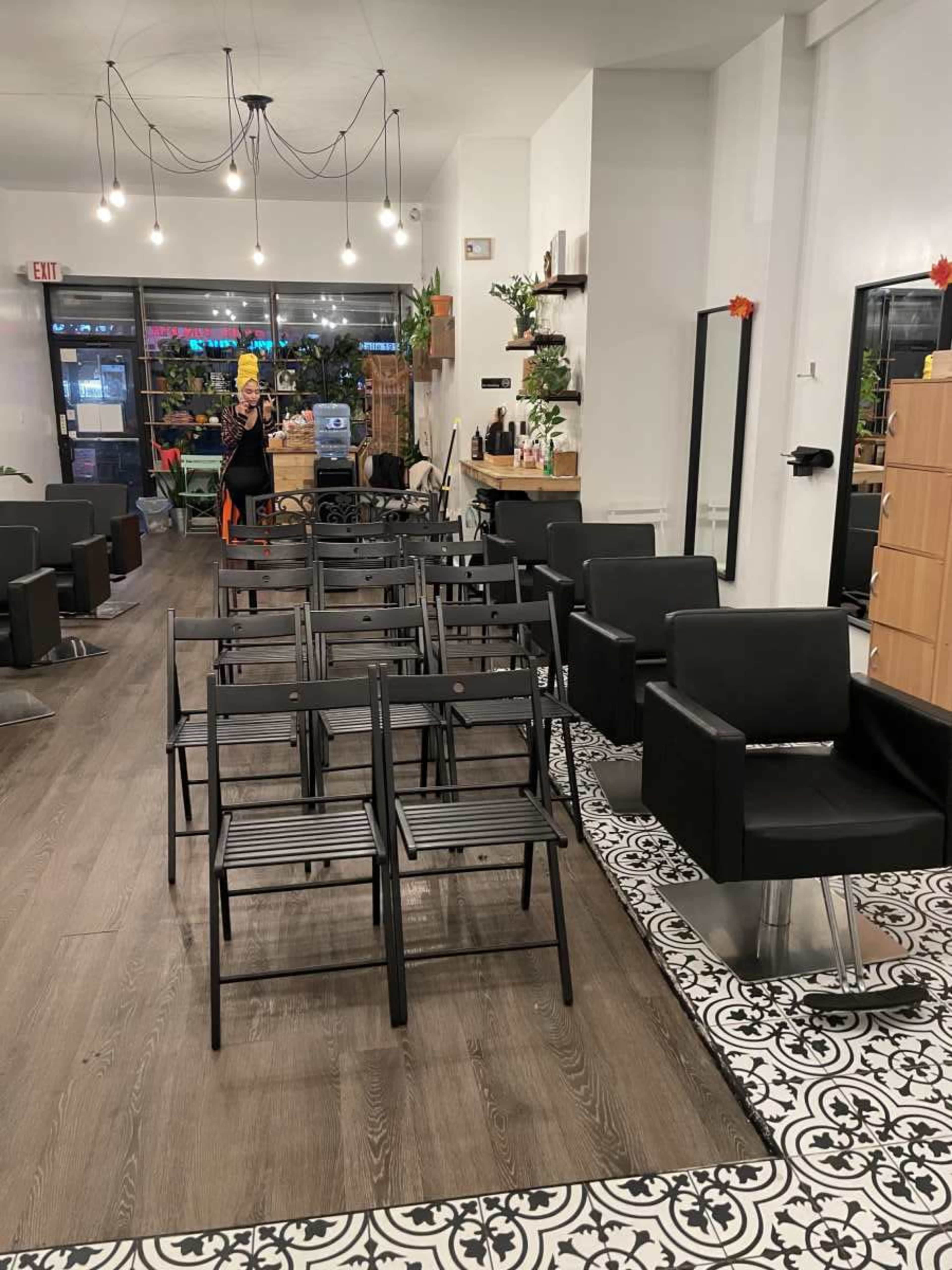 The image shows a hair salon interior with several black chairs arranged in rows on a wooden floor, and large windows providing natural light.