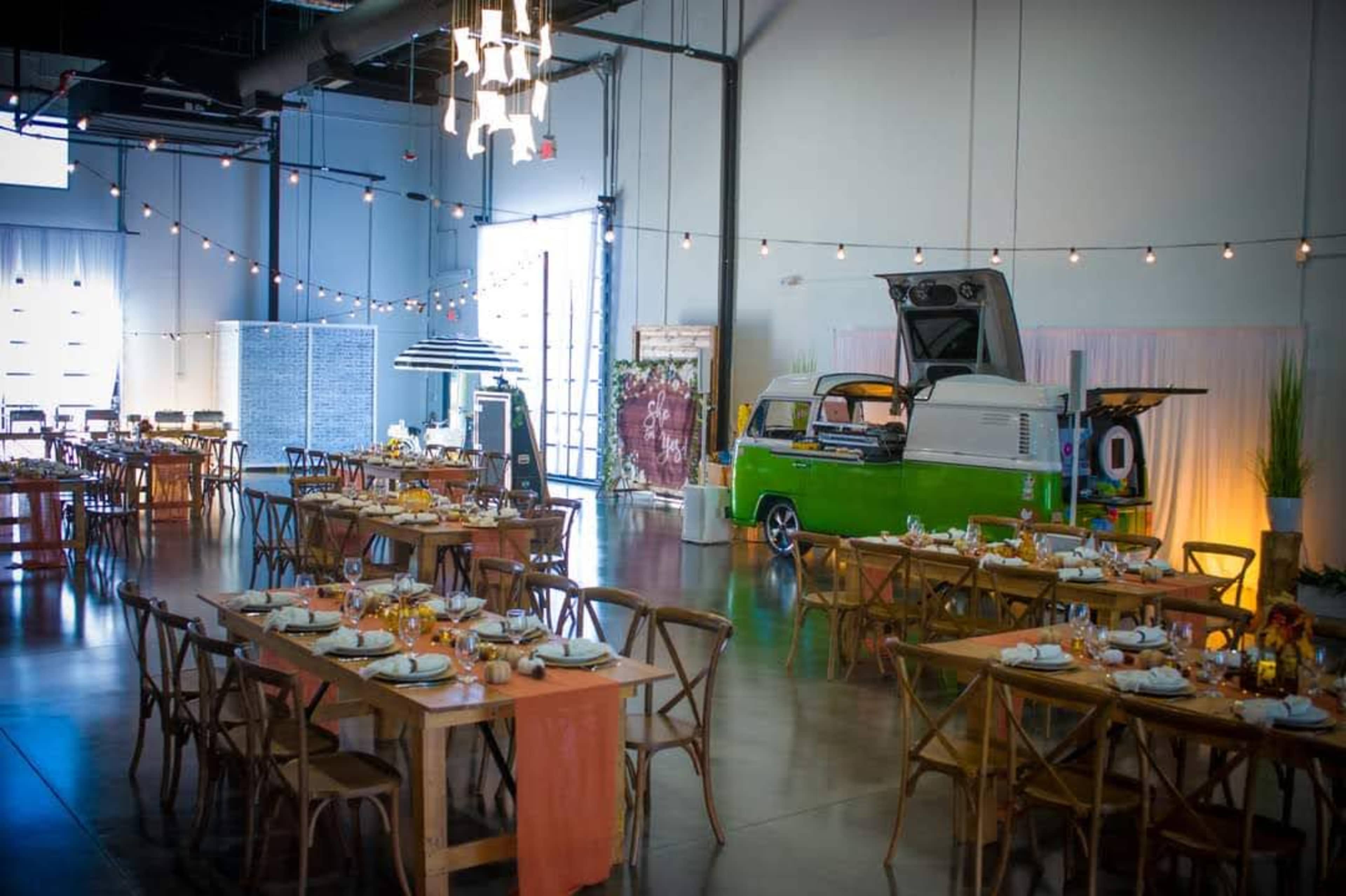 The image shows a spacious indoor event venue with wooden tables set for dining, string lights overhead, and a vintage green food truck parked in one corner.