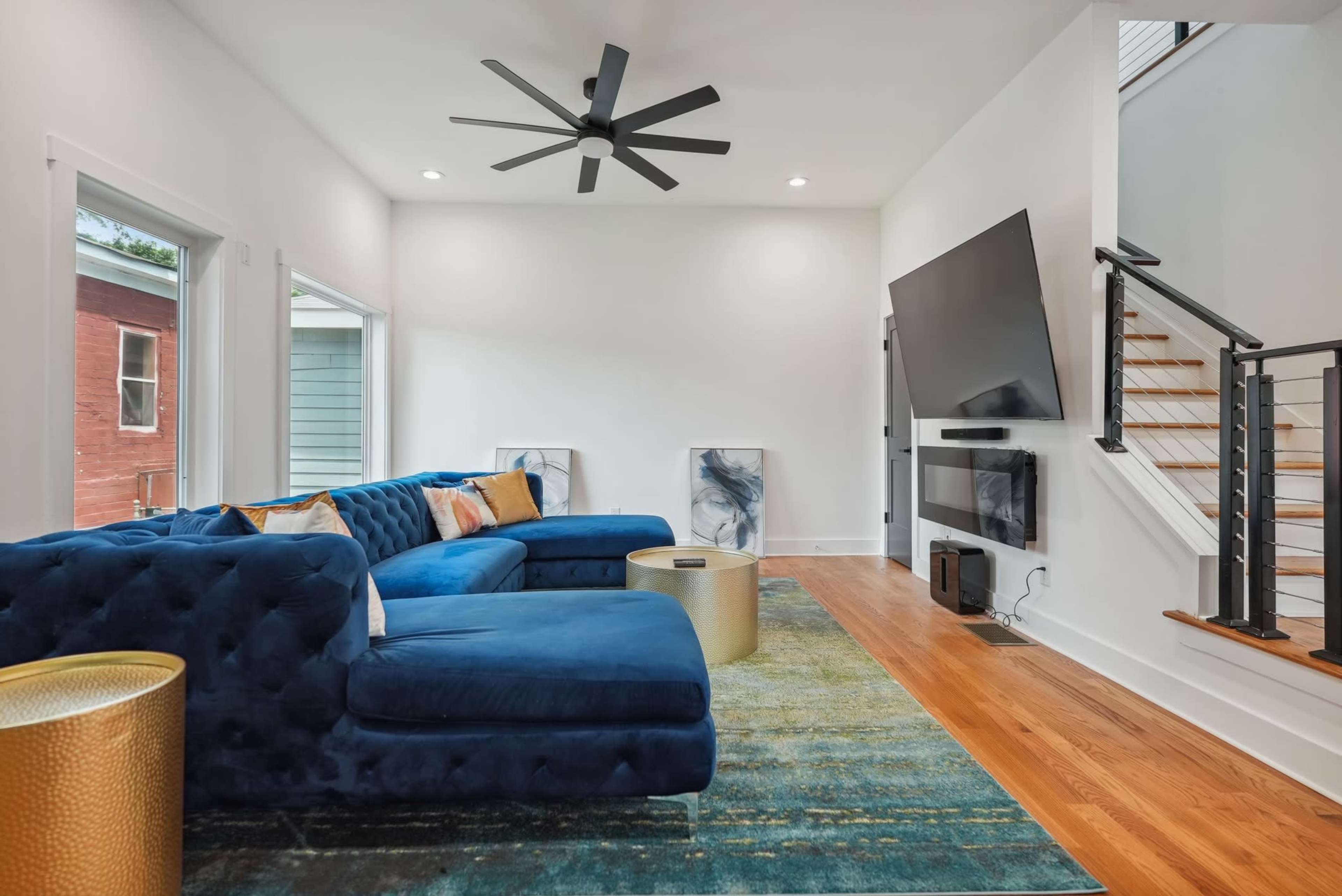 A modern living room features a blue sectional sofa, a large flat-screen TV mounted on the wall, and a staircase with a wood railing.