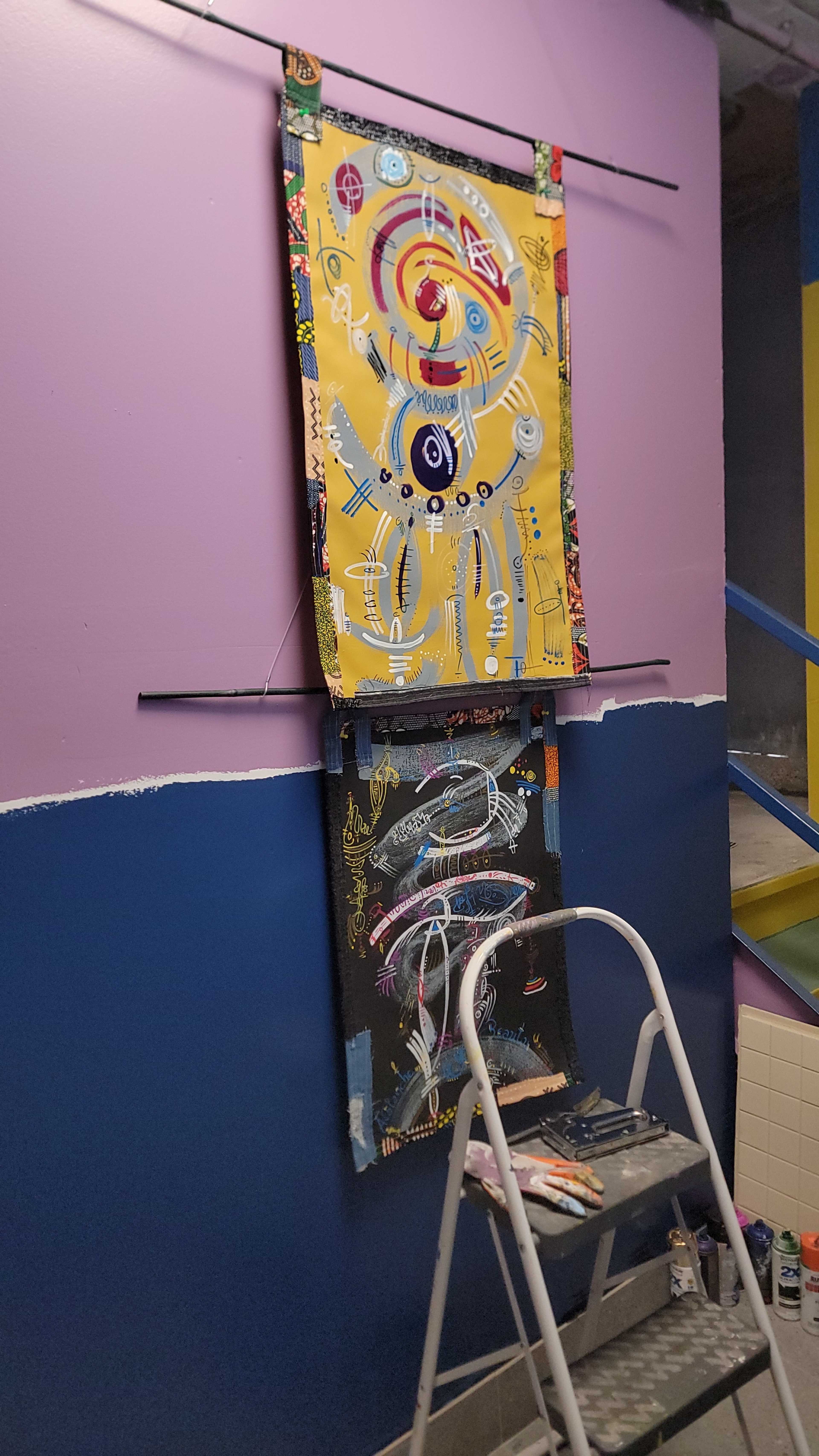 The image shows two colorful abstract paintings mounted on a purple and blue wall, with a step ladder and art supplies placed nearby.