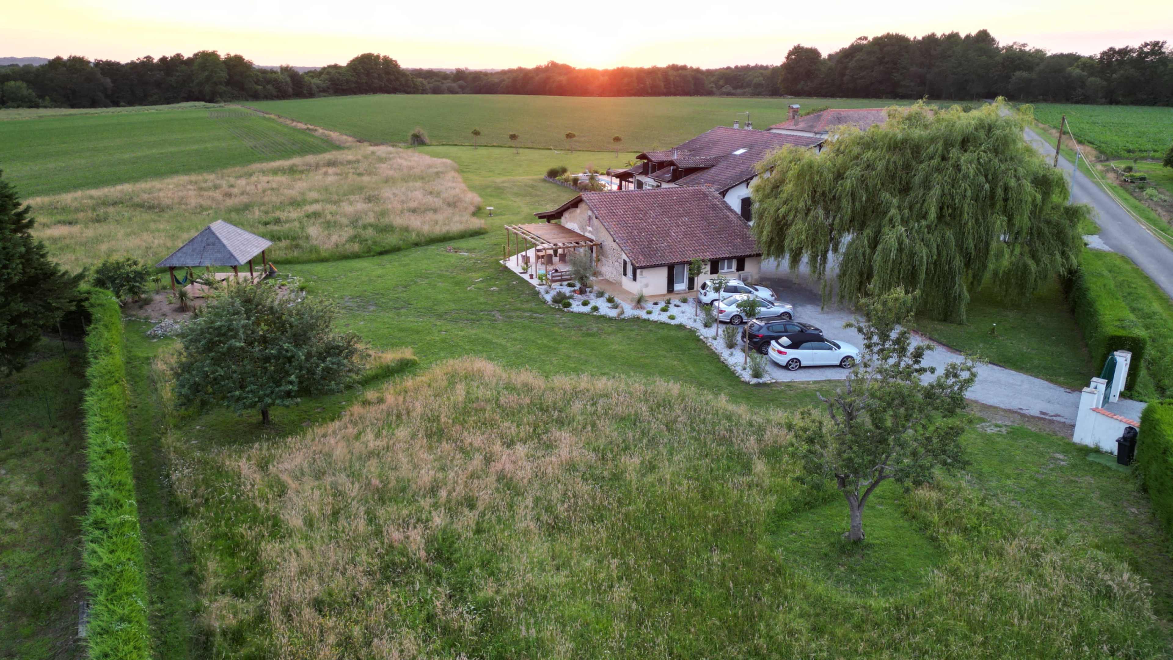 A spacious countryside property with a house, several vehicles parked, and a gazebo amidst grassy fields at sunset.