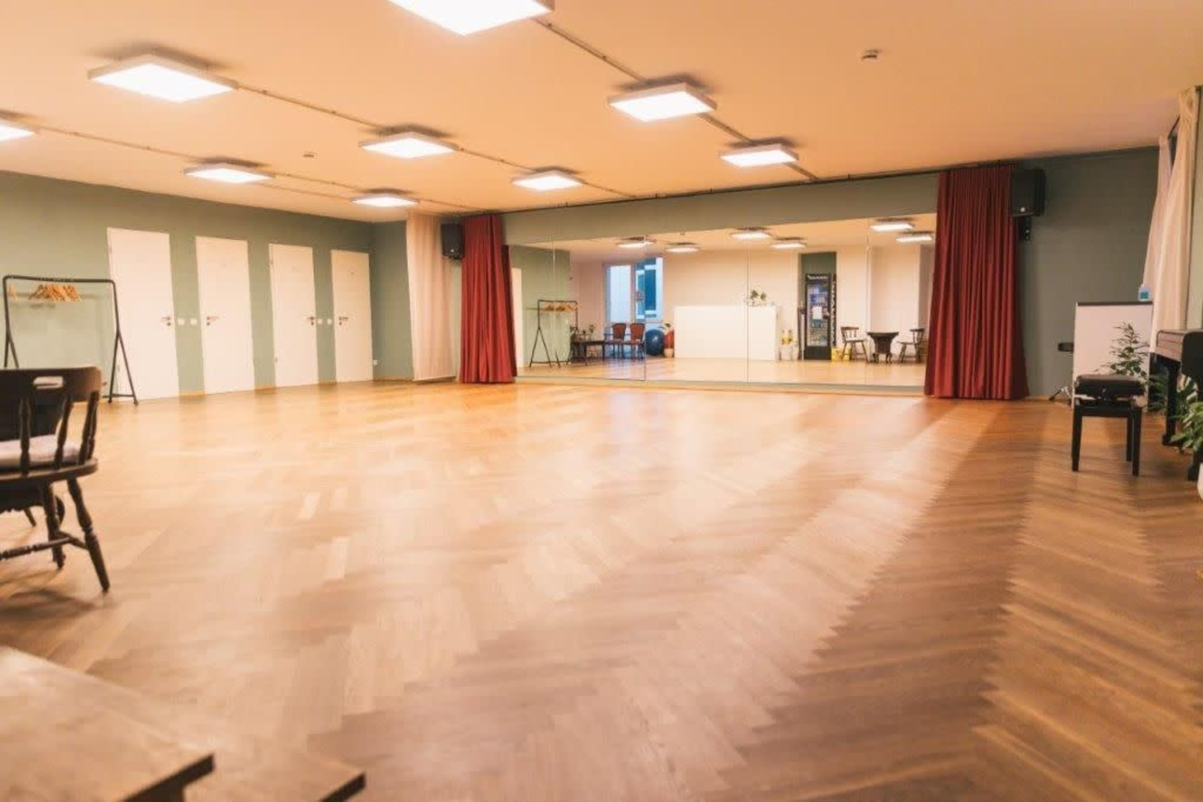 The image shows a spacious, empty room with wooden floors, a large mirror on one wall, and several doors along another wall.