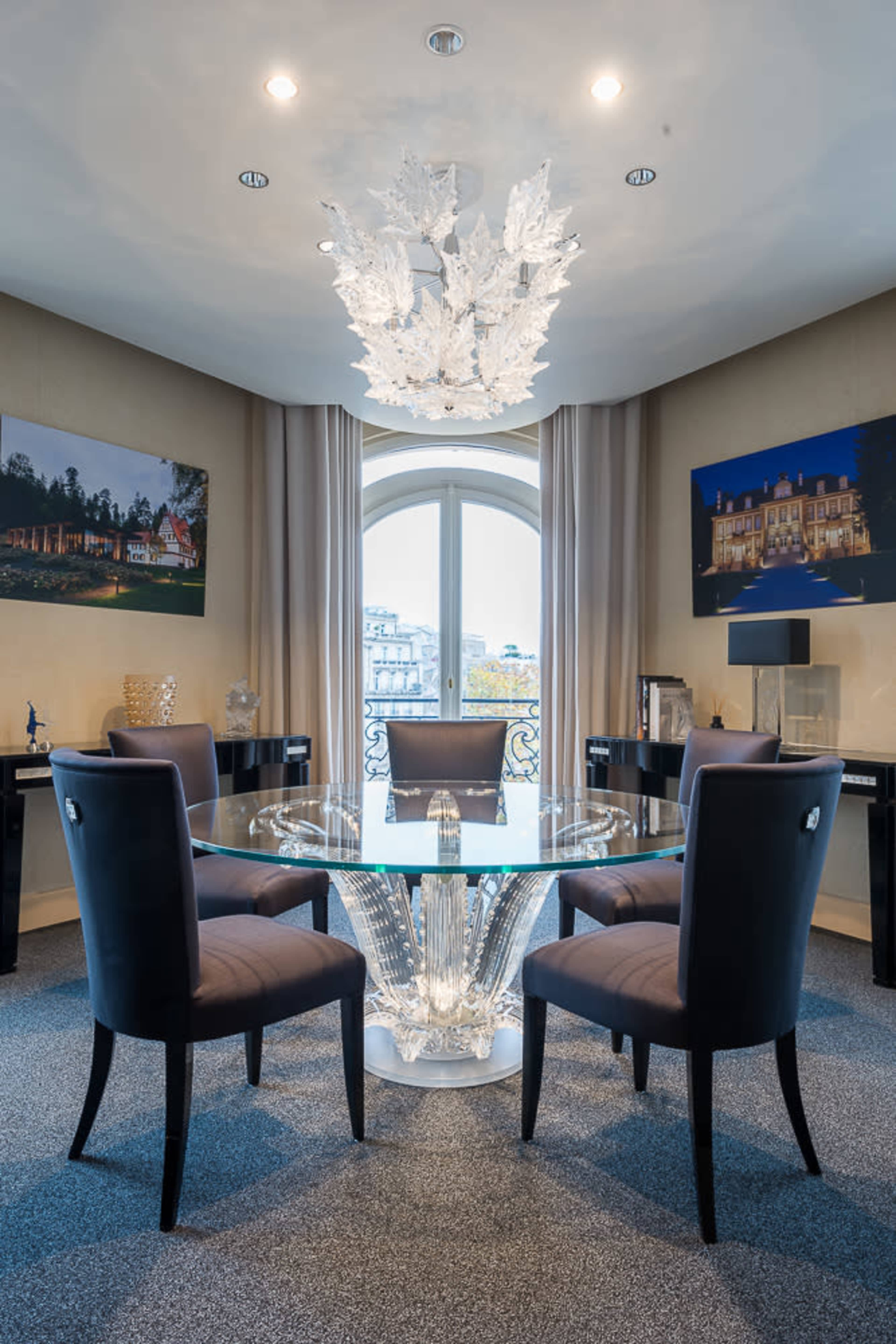 A modern dining room featuring a circular glass table with an ornate base, surrounded by four upholstered chairs, and illuminated by a decorative chandelier.