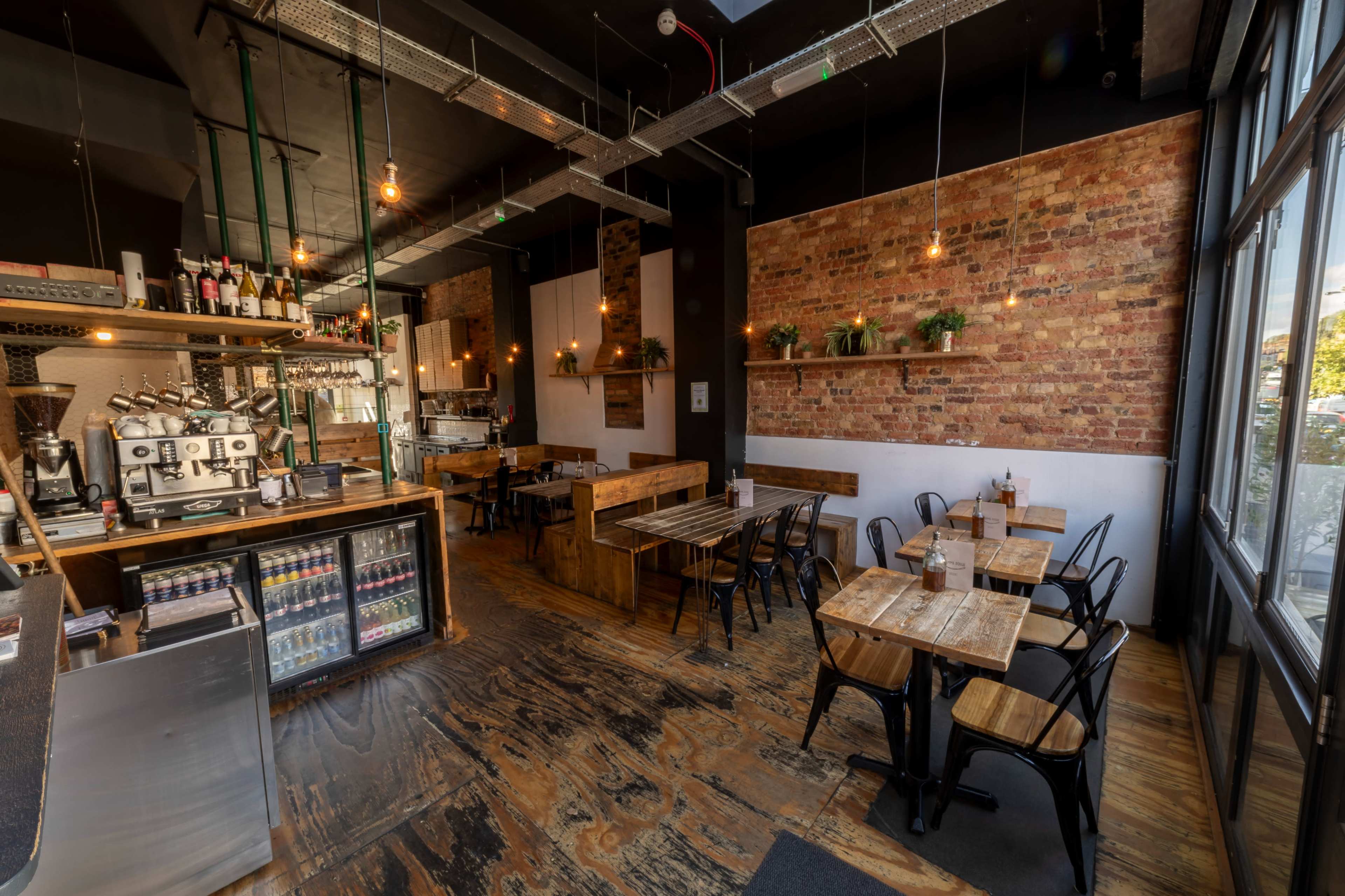 The image shows a modern café interior with exposed brick walls, wooden tables, and a coffee counter featuring machines and drinks.