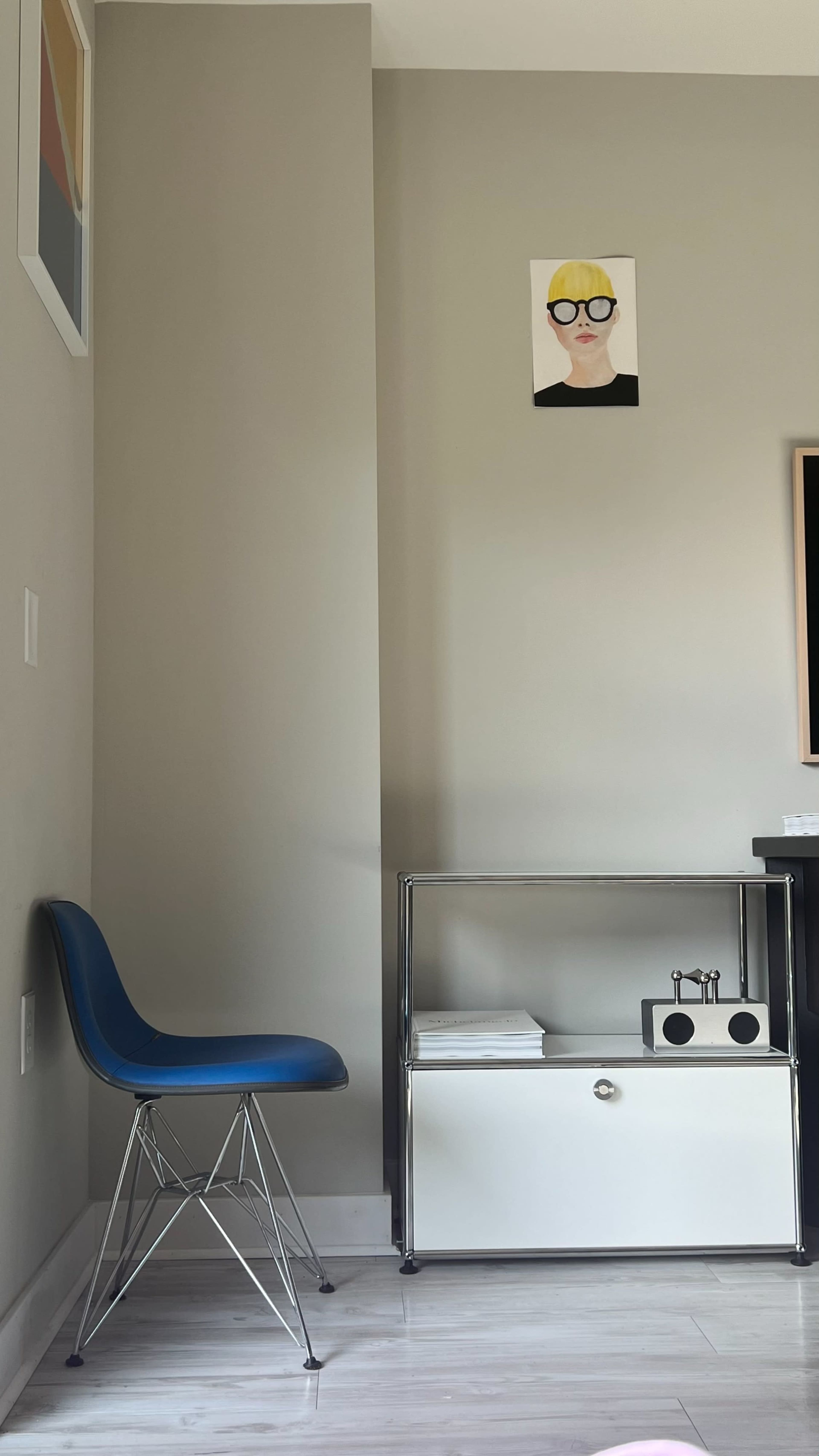 A blue chair is placed next to a white shelf with a black and white decorative piece against a light gray wall, featuring a portrait hanging above.